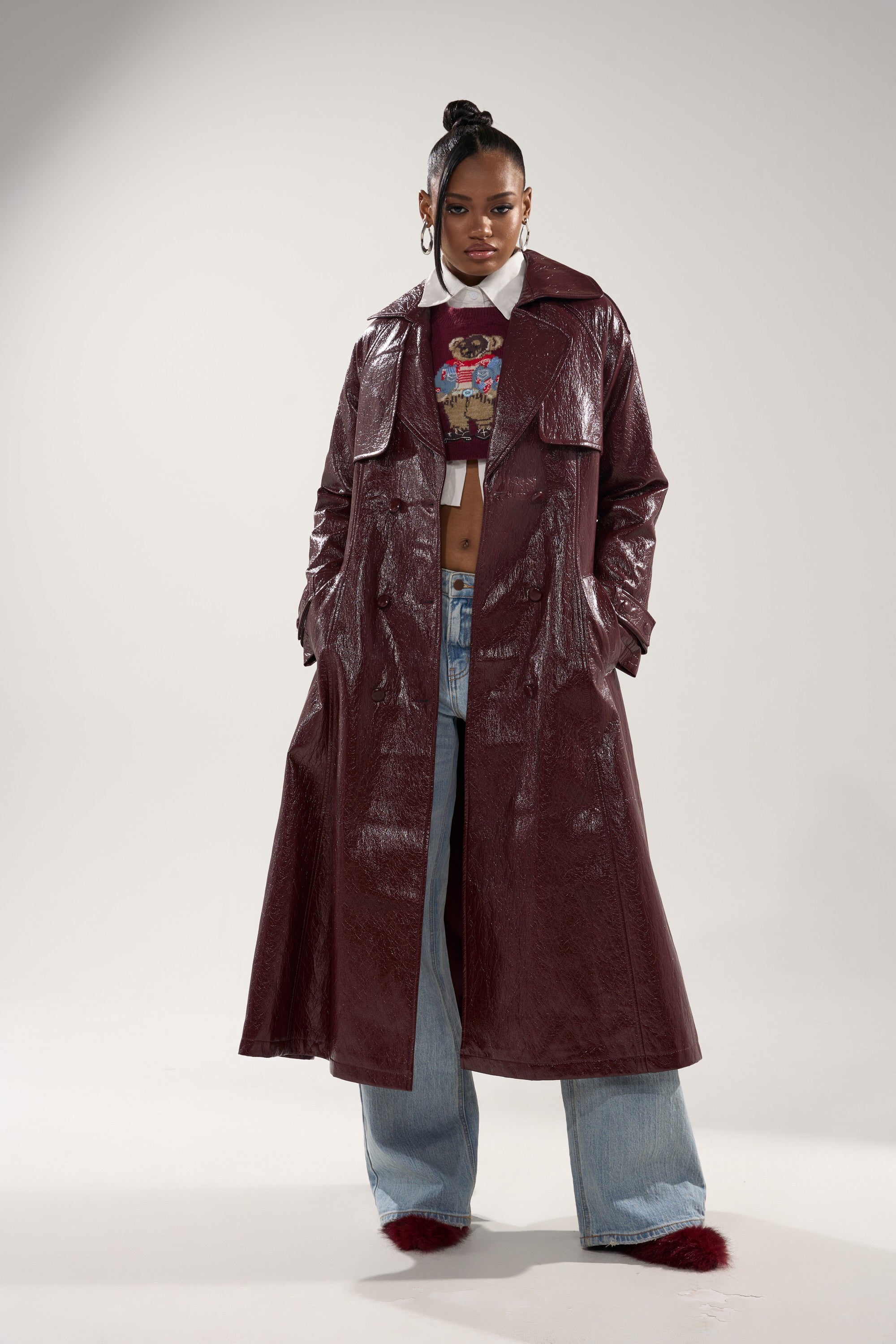 A woman in a studio models the WOKE UP IN PARIS FAUX LEATHER TRENCH, a shiny maroon longline coat, layered over a cartoon print crop top, wide-leg blue jeans, and red furry shoes. Her hair is styled in a high bun. 1