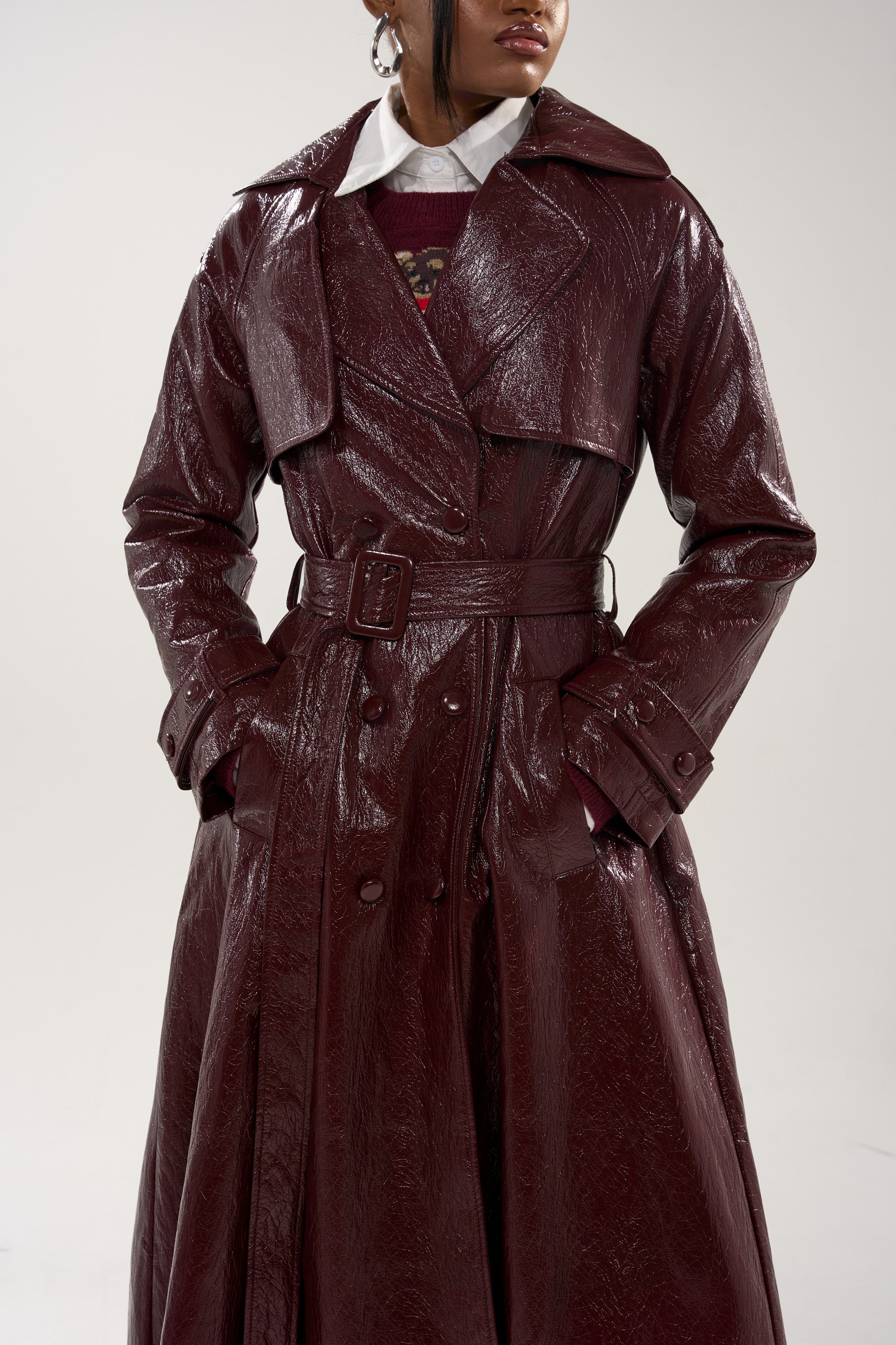 Someone wearing the WOKE UP IN PARIS FAUX LEATHER TRENCH, a dark burgundy, shiny coat with a longline silhouette and wide collar, layered over a white collared shirt, hands in pockets, head partially out of frame. 9