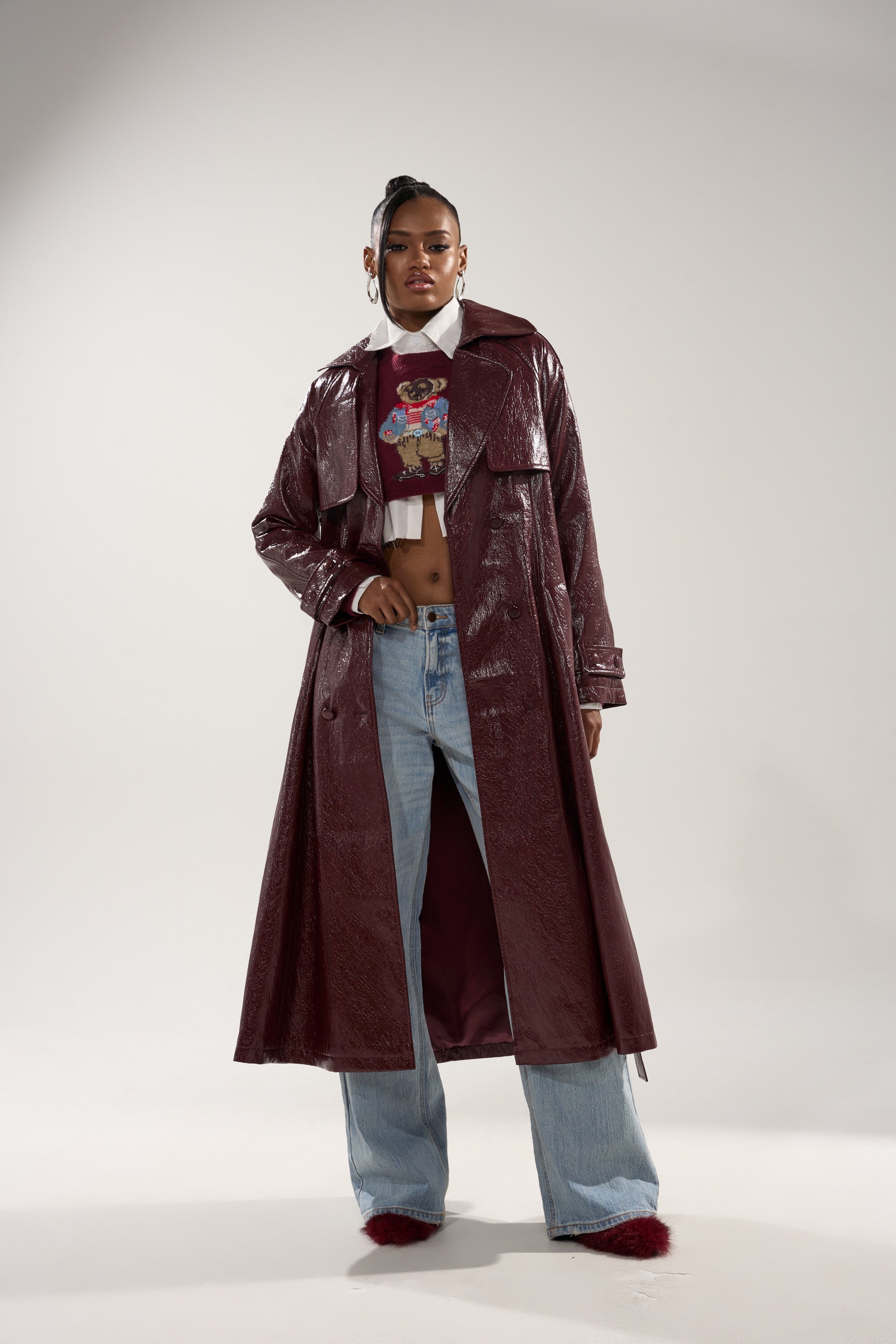 A woman stands confidently in the WOKE UP IN PARIS FAUX LEATHER TRENCH, layered over a cropped graphic top, light blue wide-leg jeans, and maroon fur-trimmed shoes against a plain light background. 2