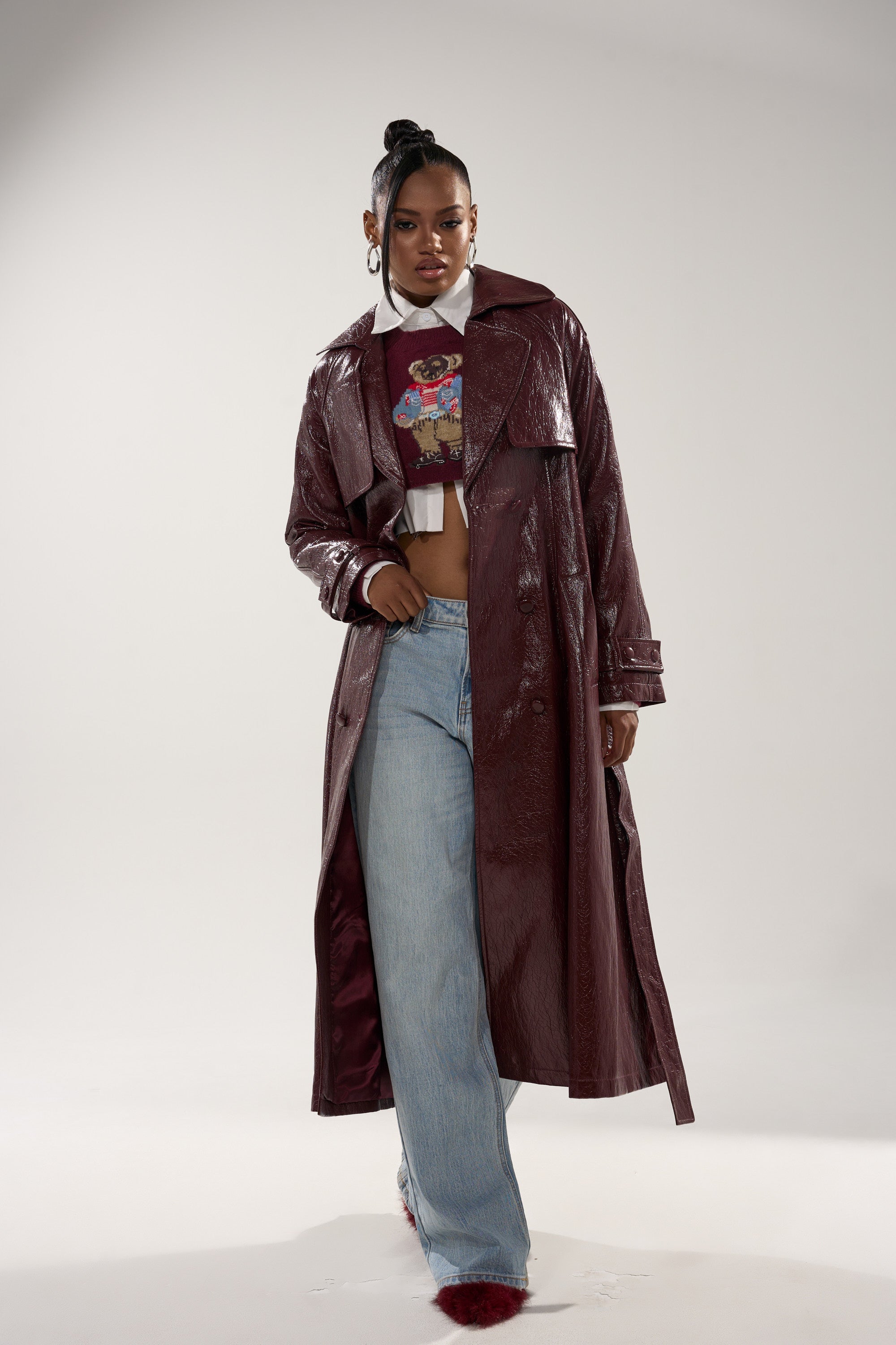 A woman stands confidently in the WOKE UP IN PARIS FAUX LEATHER TRENCH over a cropped cartoon-print sweater, a white collared shirt, and light blue wide-leg jeans against a plain light background. 3