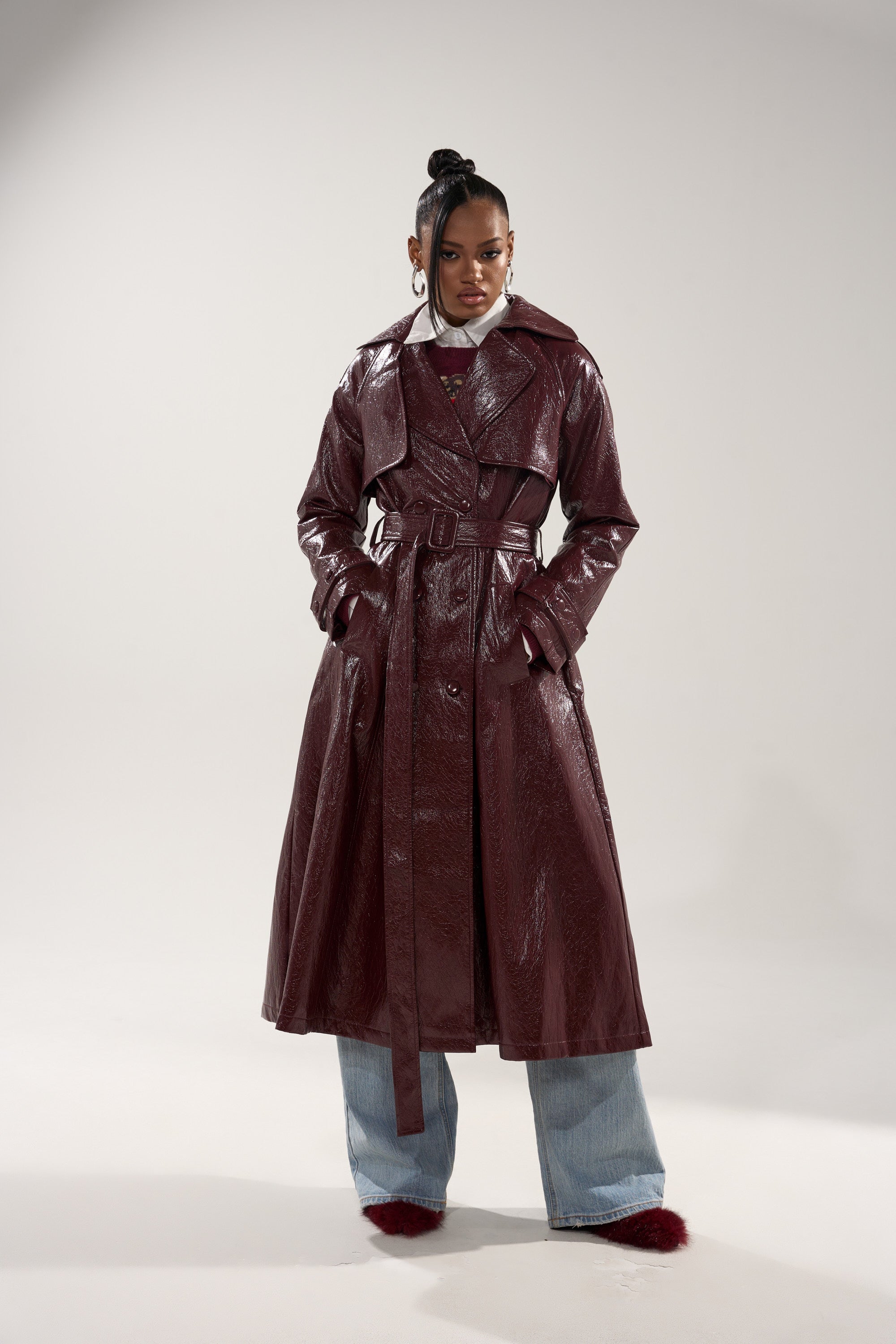 A woman confidently wears the WOKE UP IN PARIS FAUX LEATHER TRENCH in glossy maroon over light blue wide-leg jeans and furry maroon shoes, her hair in a high bun with large hoop earrings. 0