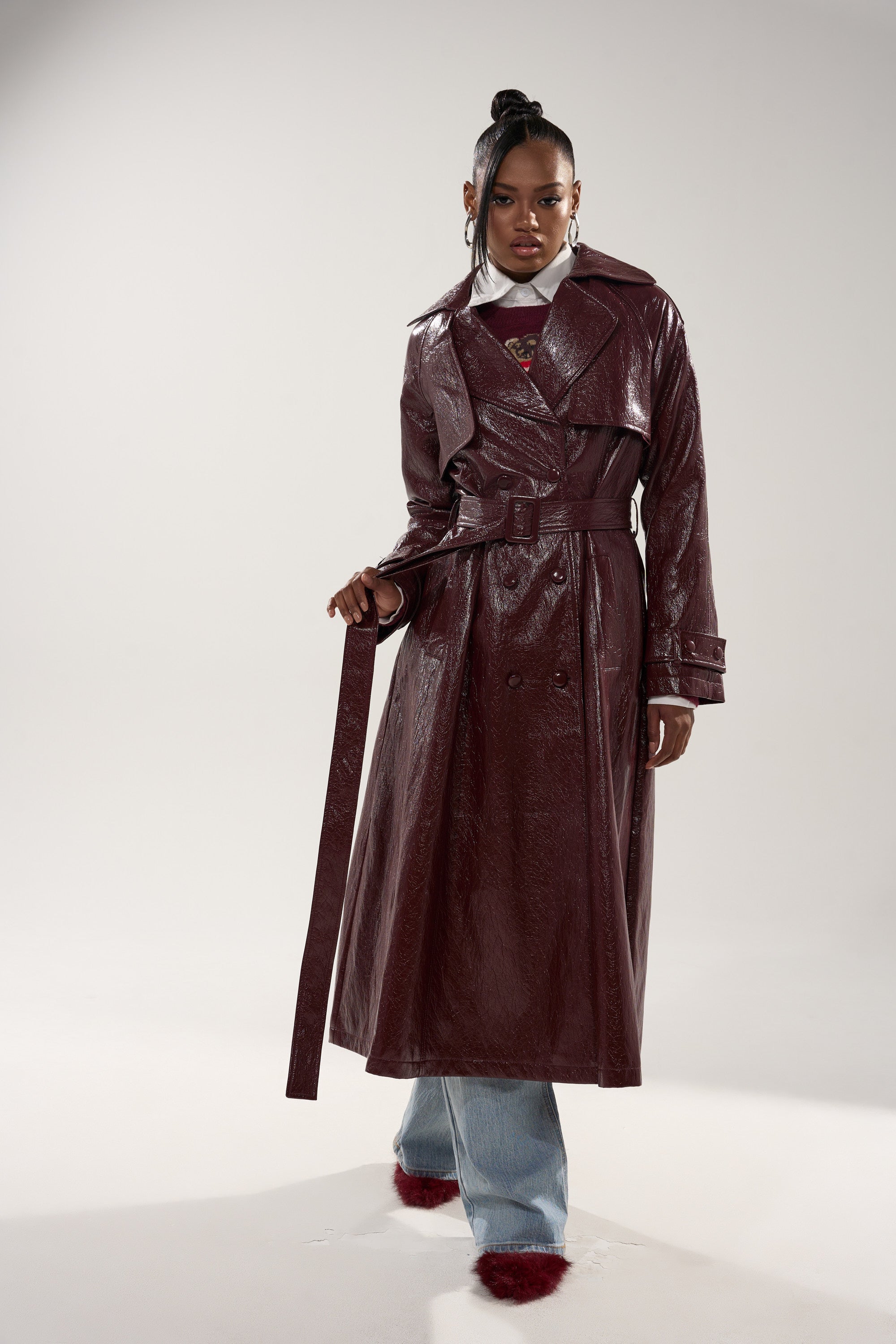 A woman models the WOKE UP IN PARIS FAUX LEATHER TRENCH in shiny dark burgundy, belted over a white top and blue jeans. She stands confidently with her hair up and statement earrings, highlighting the coat’s longline silhouette. 4