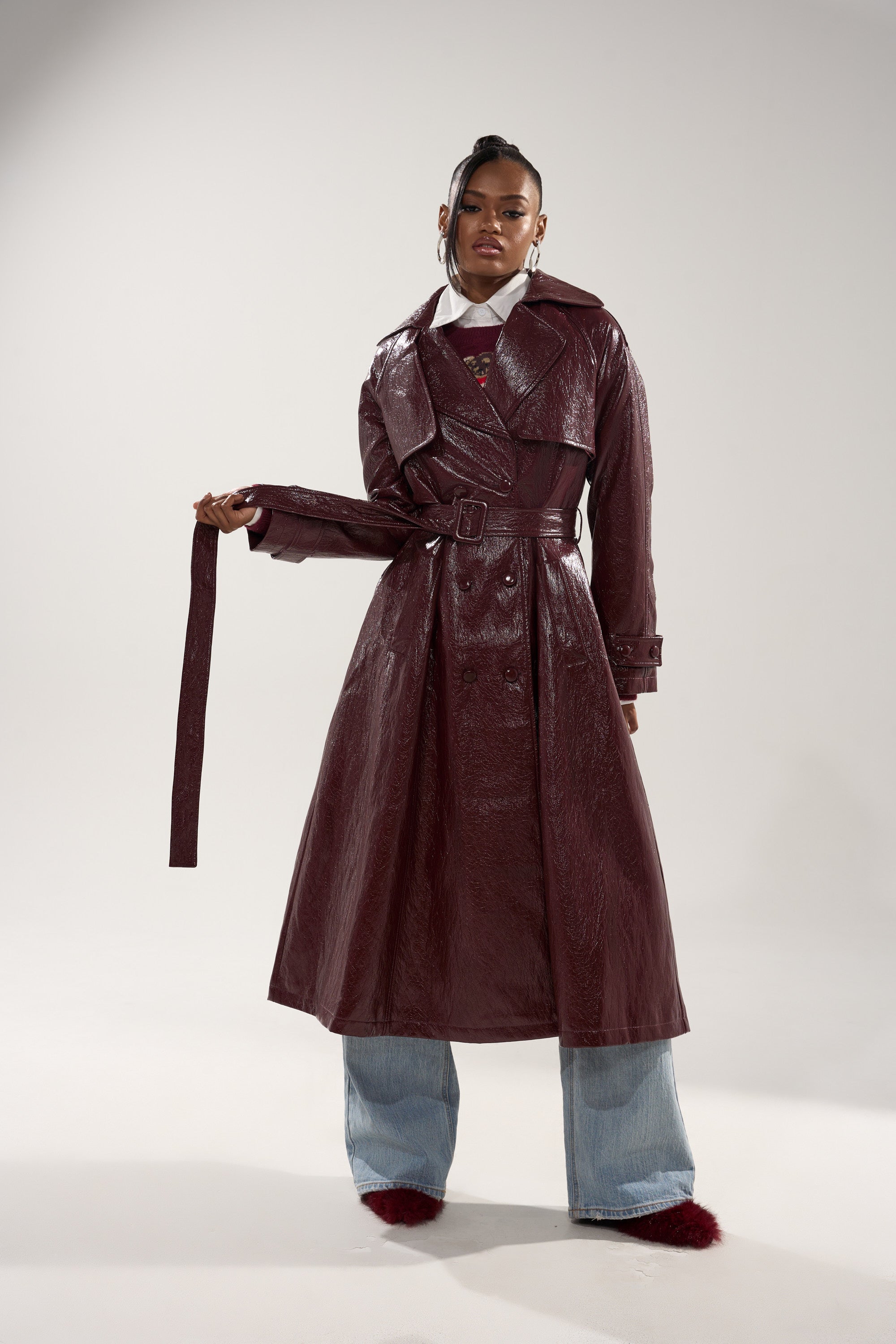 A woman poses in the WOKE UP IN PARIS FAUX LEATHER TRENCH with a matching belt, paired with wide-leg light blue jeans and red fuzzy shoes, against a plain light background. 5