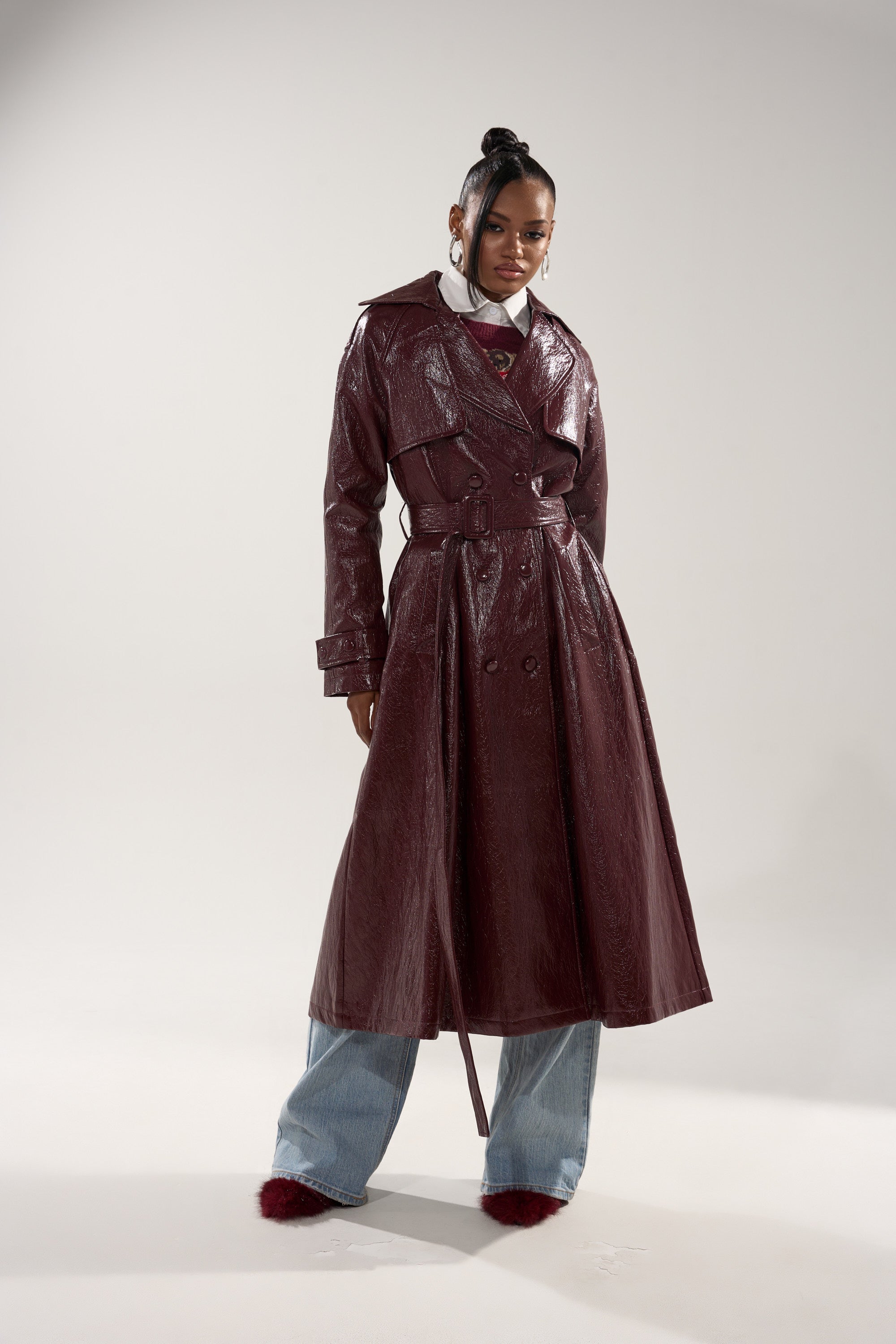A woman confidently poses against a plain background wearing the WOKE UP IN PARIS FAUX LEATHER TRENCH—a shiny maroon longline coat—styled with wide-legged blue jeans, red shoes, and her hair in a high bun. 6