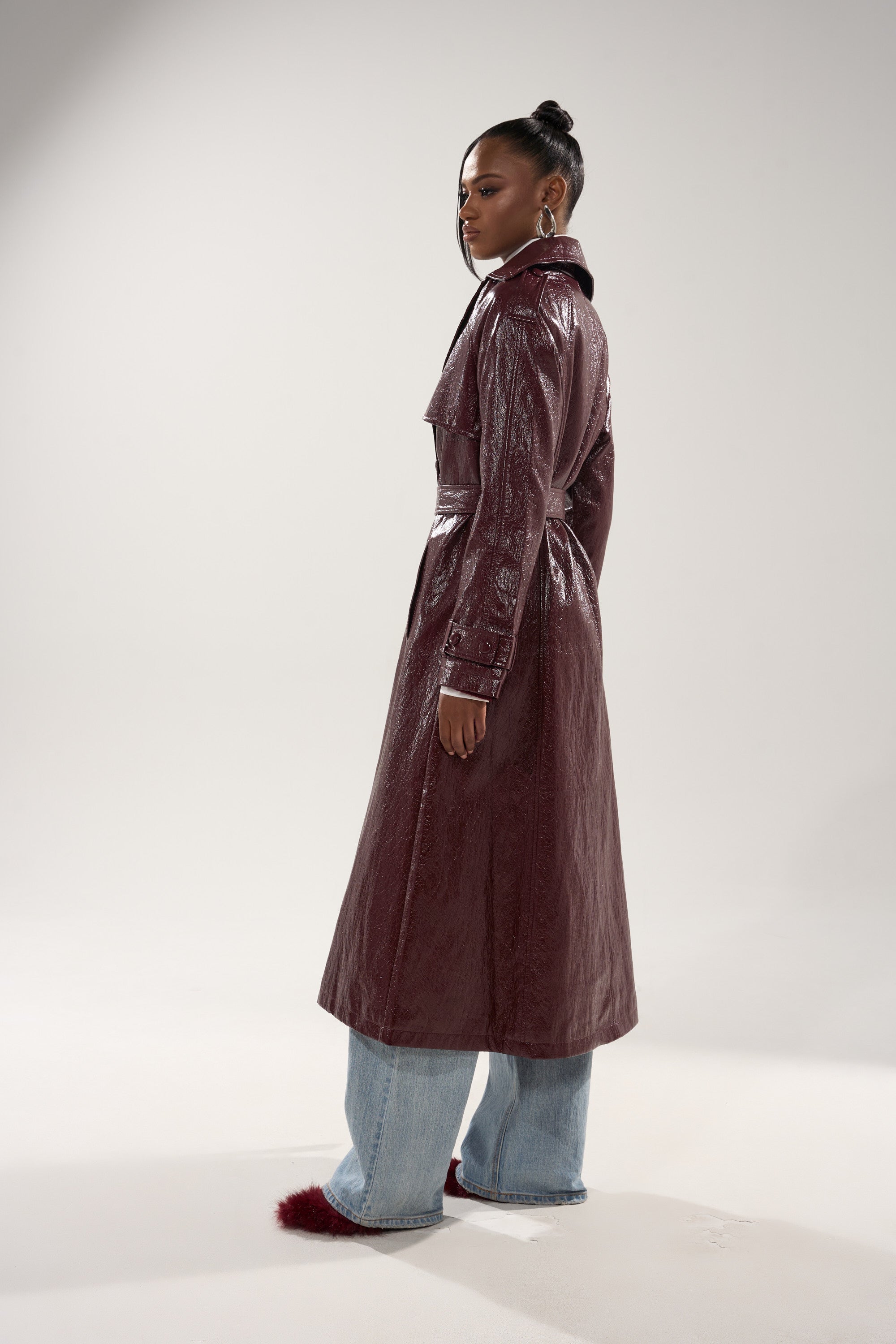 A woman looks over her shoulder, wearing the WOKE UP IN PARIS FAUX LEATHER TRENCH in shiny burgundy, paired with wide-leg light blue jeans and burgundy fluffy shoes against a plain light background. 7