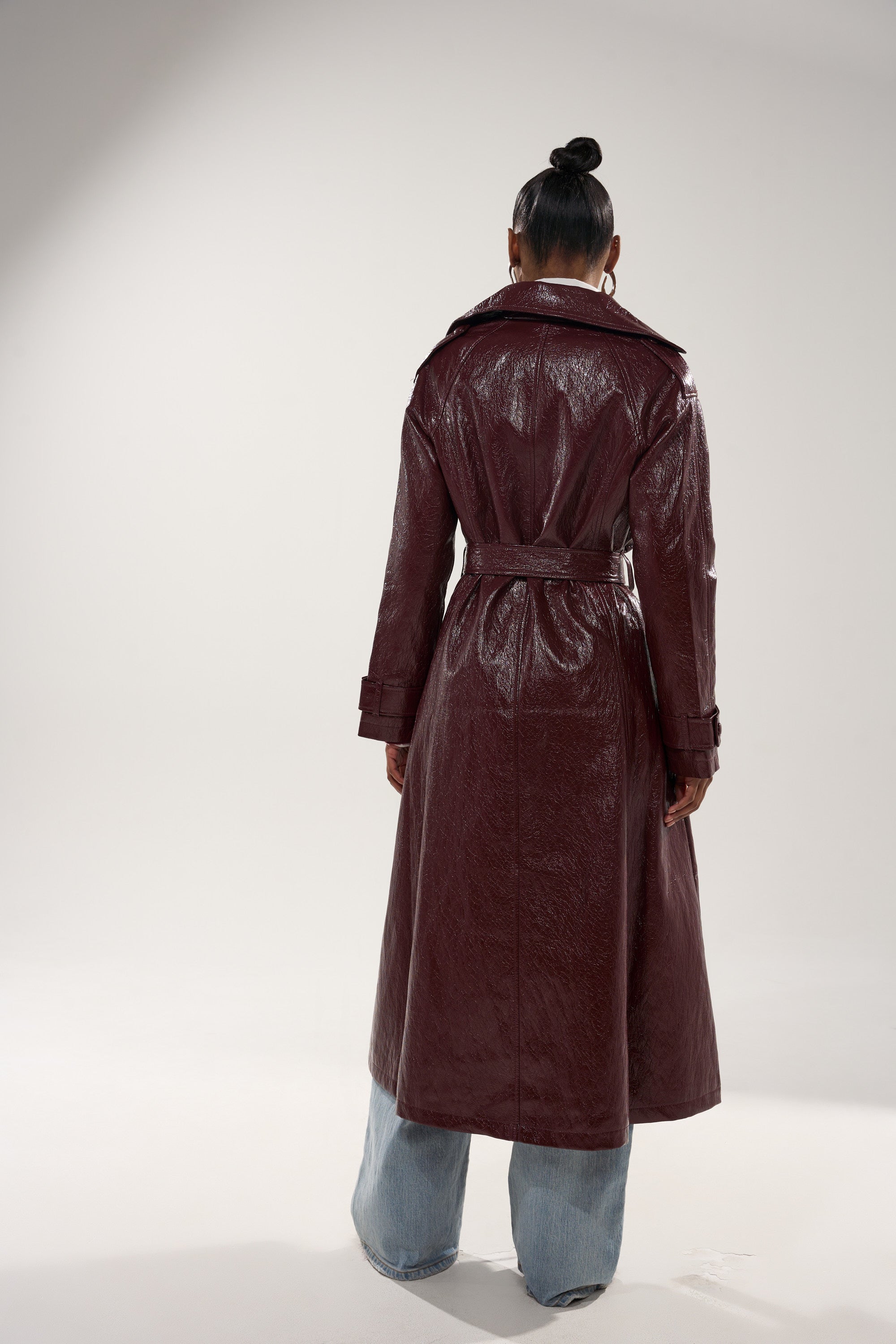 A person faces away in the WOKE UP IN PARIS FAUX LEATHER TRENCH, a shiny maroon, belted coat with a wide collar, paired with light blue jeans. The longline style complements their neat bun against a plain, neutral backdrop. 8