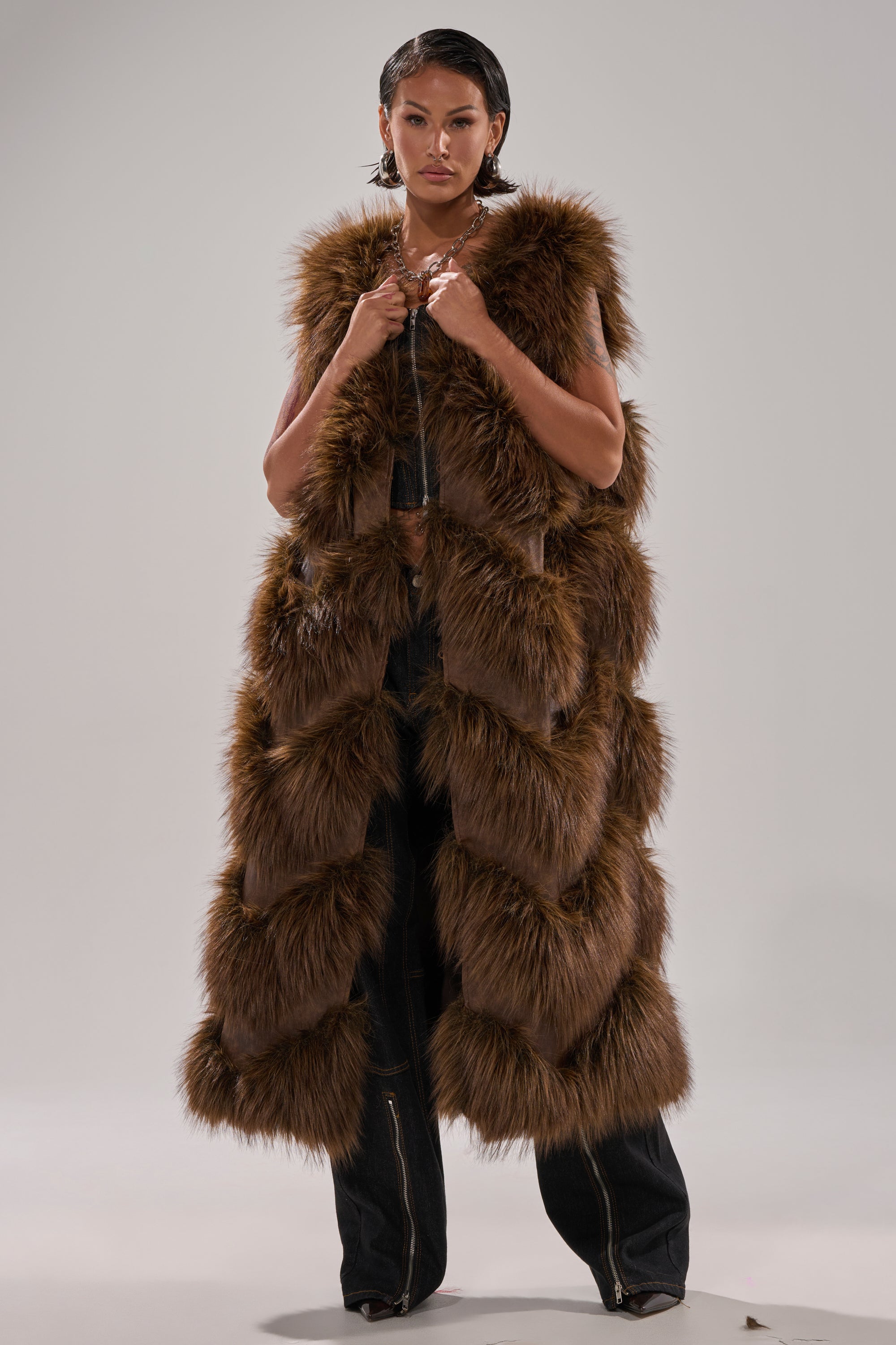 A person stands facing forward in the CAN'T BE BOTHERED PANELED FUR VEST, a longline brown faux fur piece with a layered texture, worn over dark pants against a plain, light background.