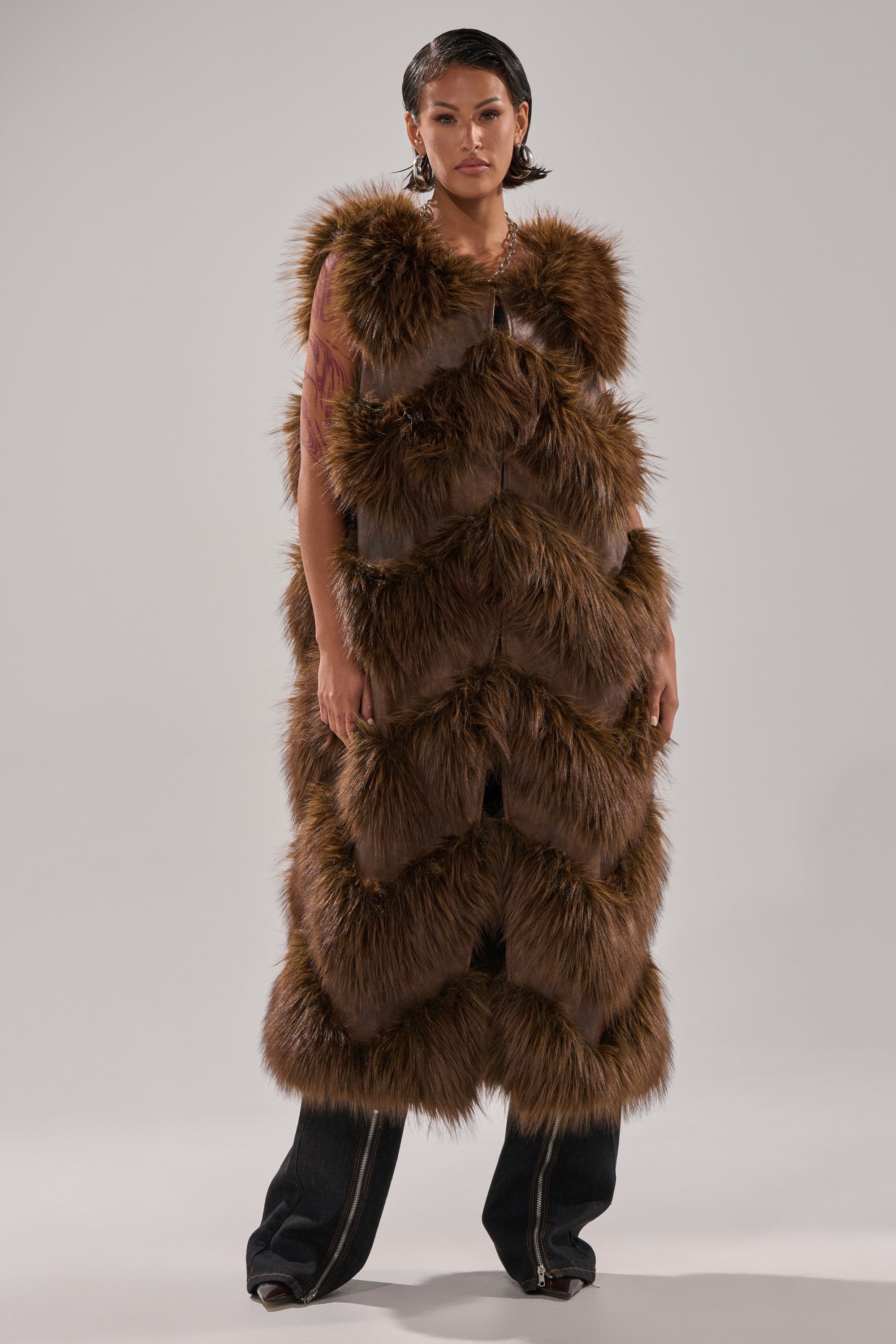 A woman faces forward in the CAN'T BE BOTHERED PANELED FUR VEST, a brown longline faux fur piece with a chevron pattern, styled over dark pants against a light plain background.
