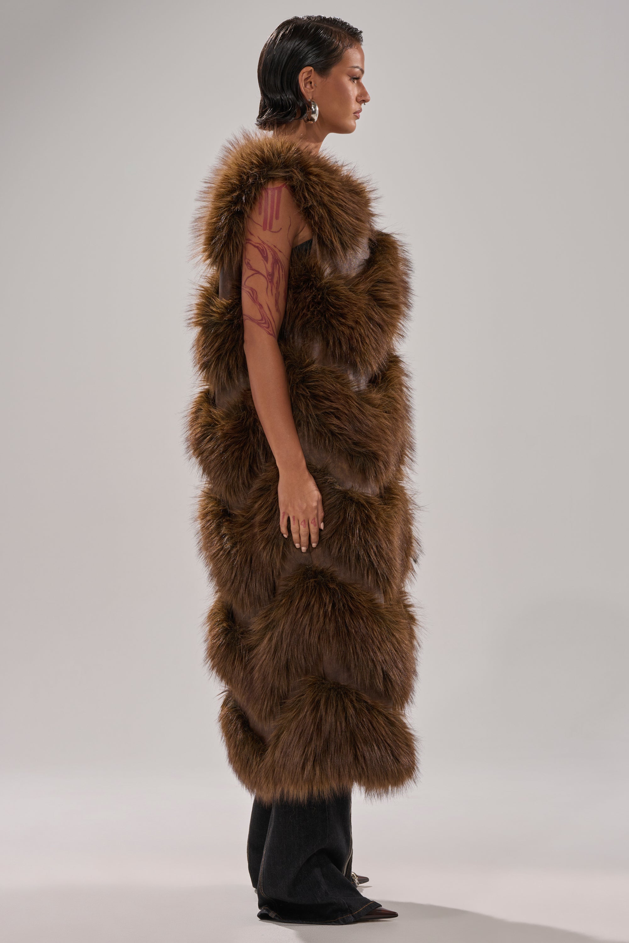 A person with short, slicked-back hair stands in profile, showcasing a tattooed arm while wearing the CAN'T BE BOTHERED PANELED FUR VEST over black pants and pointed shoes against a plain light background.
