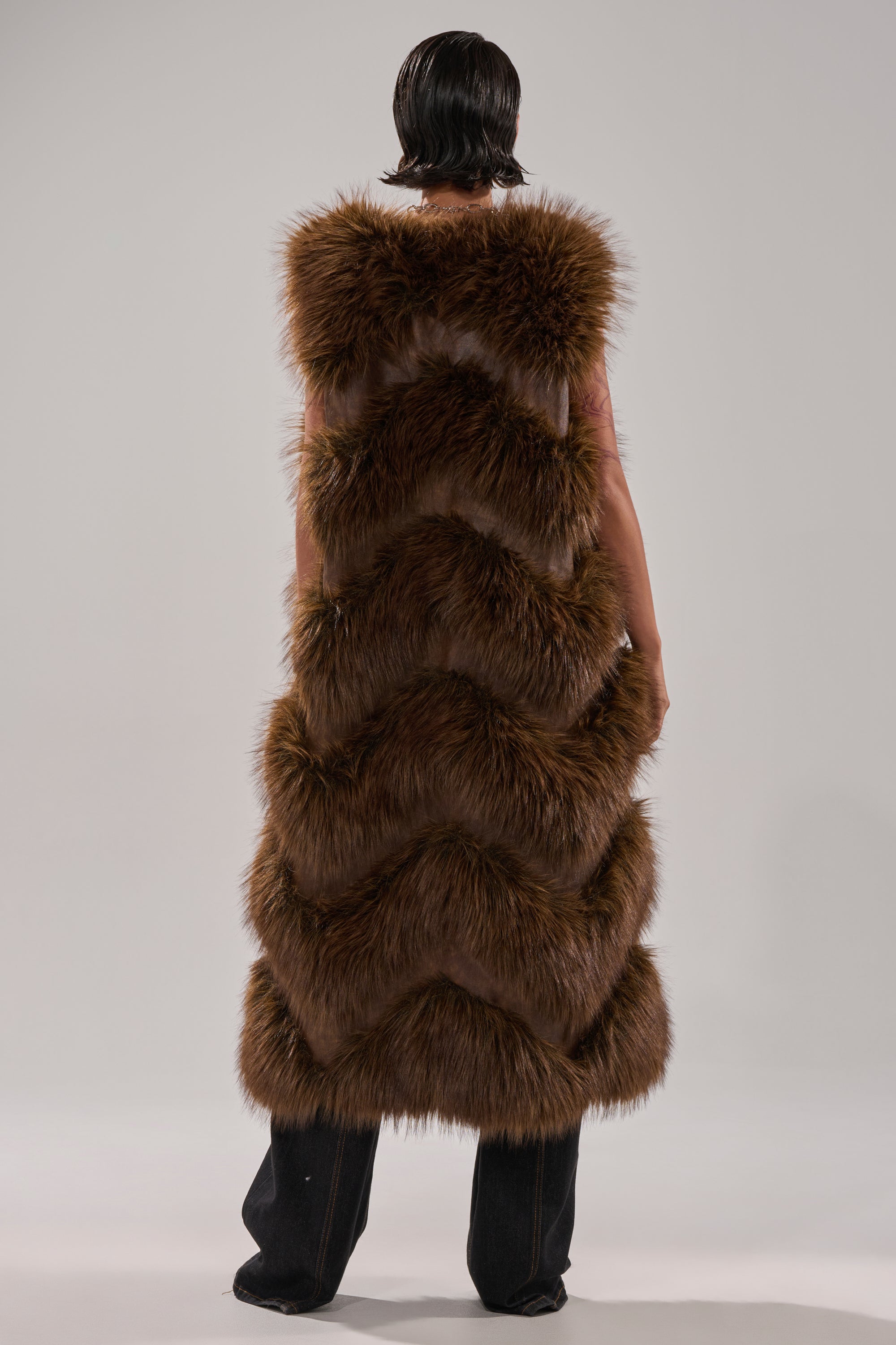 A person stands facing away in the CAN'T BE BOTHERED PANELED FUR VEST—a long, sleeveless brown faux fur vest with a chevron pattern—paired with dark wide-leg pants against a plain light gray background.