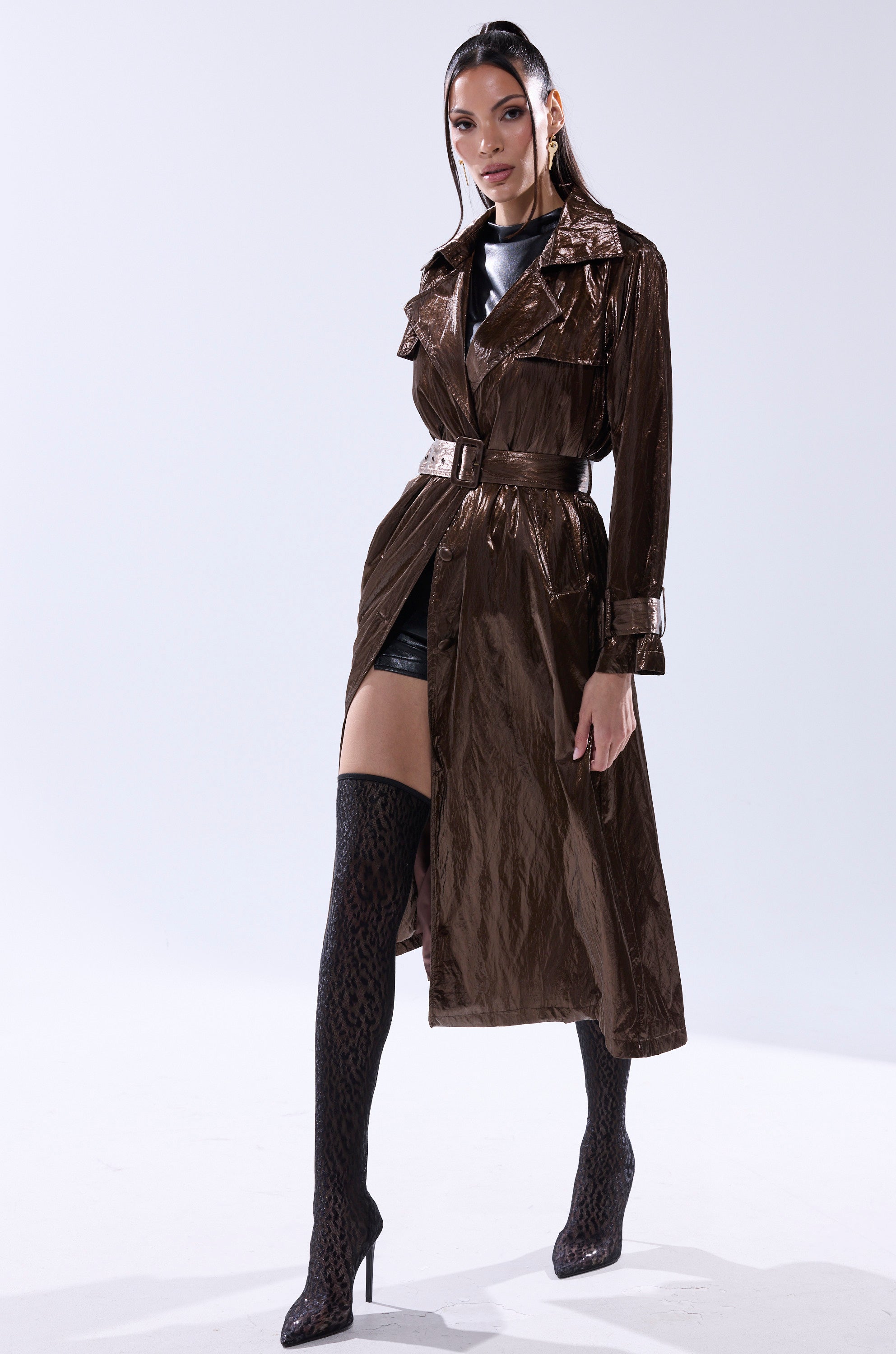 A woman poses confidently in the WORK OF ART METALLIC TRENCH, paired with a black top, black shorts, and thigh-high patterned high-heeled boots against a plain white background. 0