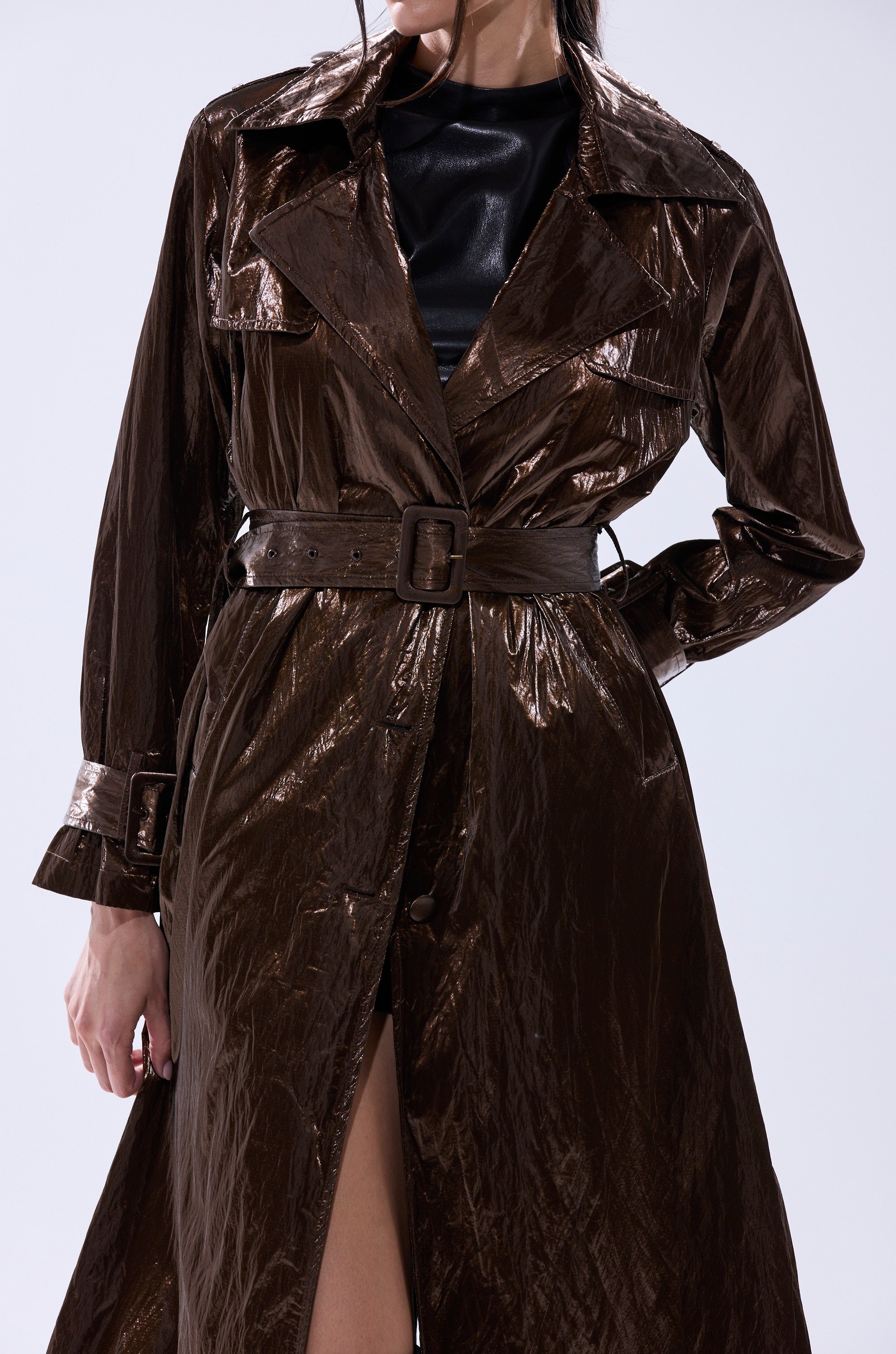 A person wears the WORK OF ART METALLIC TRENCH in shiny dark brown over a black top, with the image cropped above the nose and one leg slightly exposed through the coat’s front opening. 9