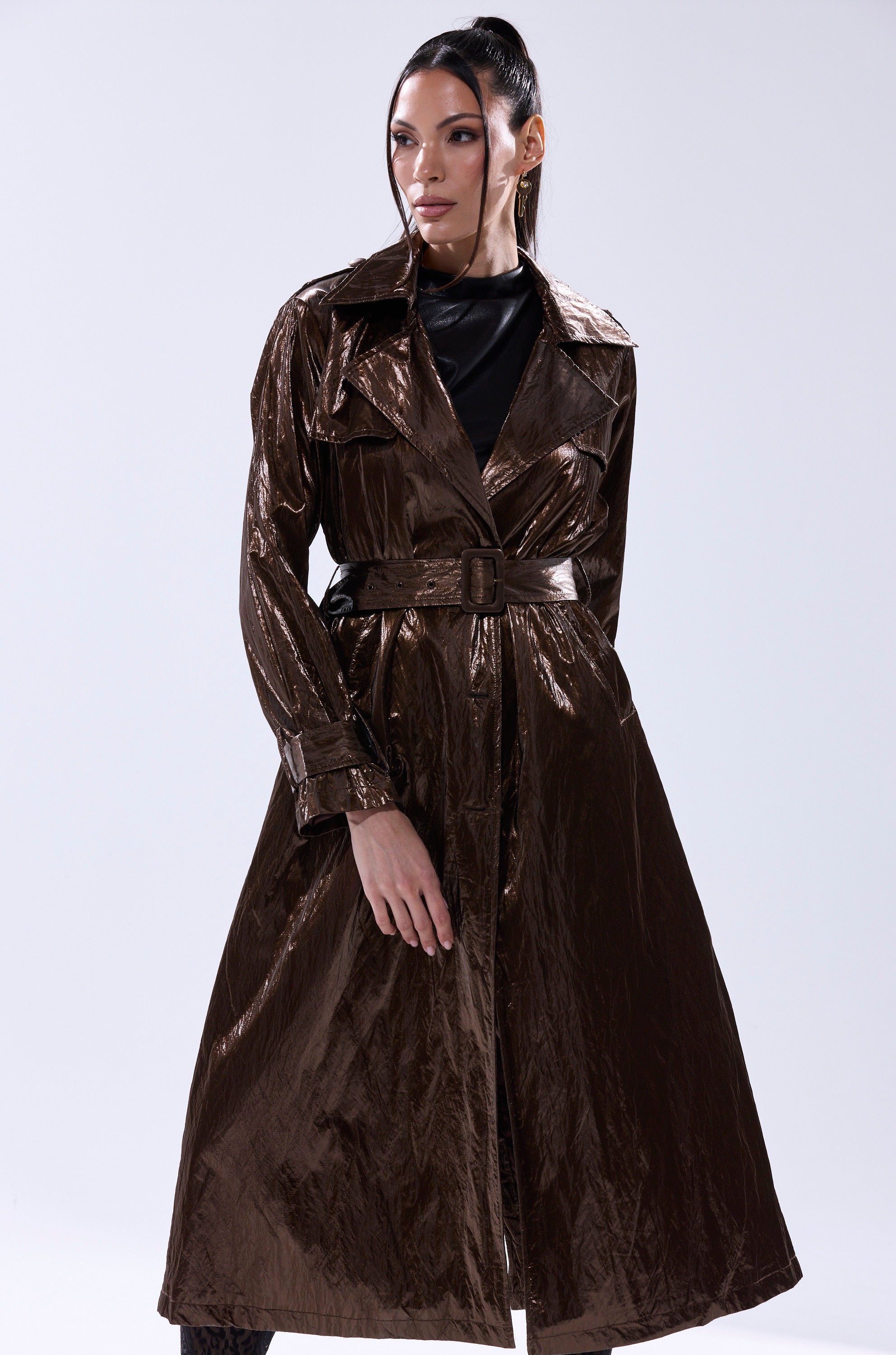 A woman models the WORK OF ART METALLIC TRENCH, a shiny bronze longline coat with a belted waist, over a black top, set against a plain light background. 1