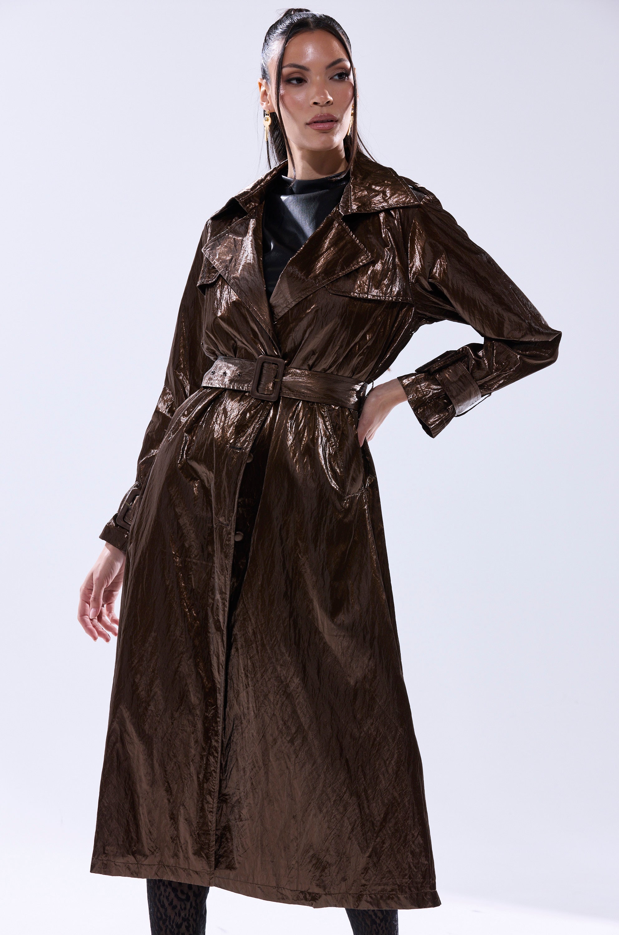 A woman stands confidently in the WORK OF ART METALLIC TRENCH, featuring a wide collar and long sleeves, paired with dark patterned tights—a true statement piece against a plain light background. 2