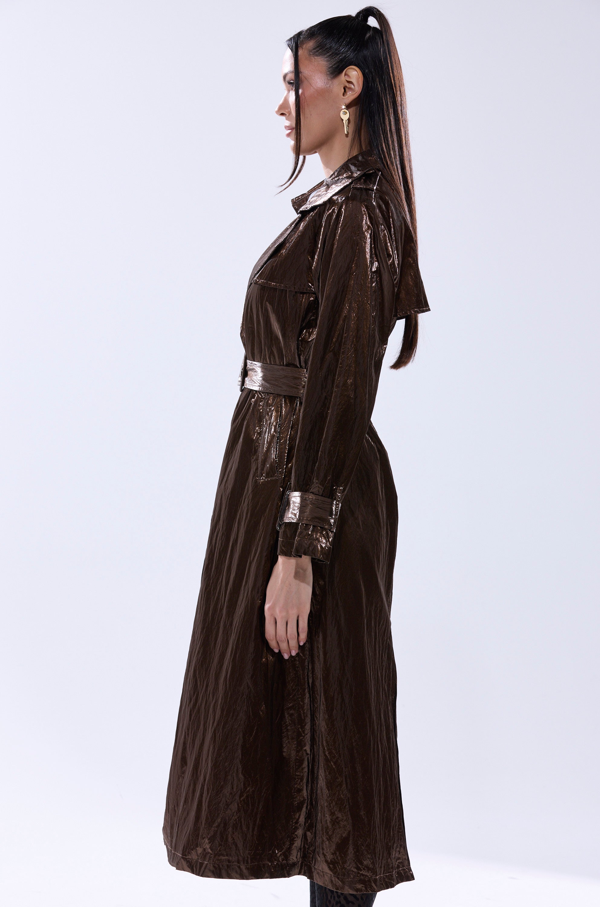 A woman in profile wears the WORK OF ART METALLIC TRENCH, a long belted coat with buttoned cuffs. Her dark hair is tied in a high ponytail, standing against a plain light background. 5