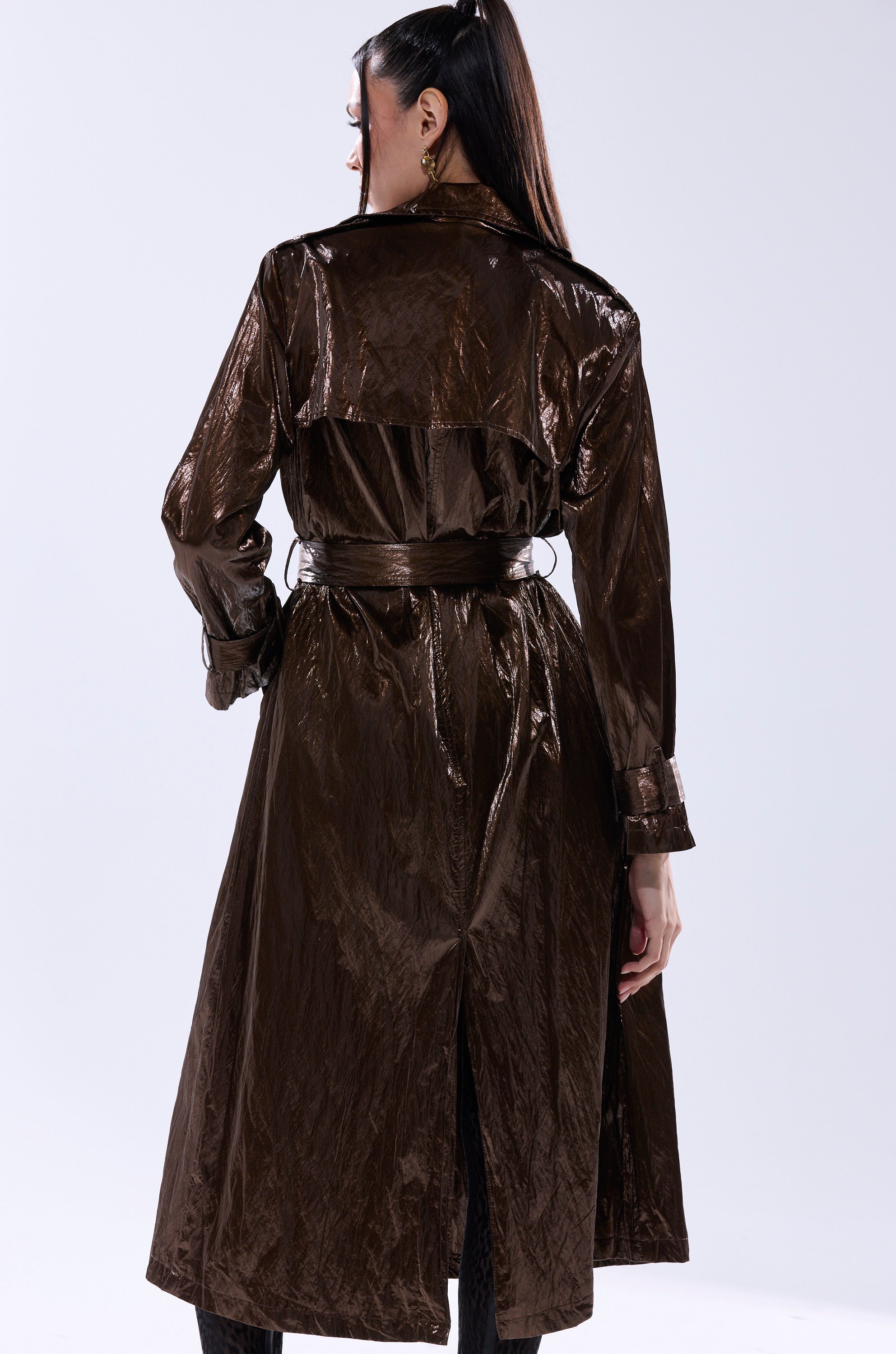 A woman faces away, wearing the WORK OF ART METALLIC TRENCH—a longline bronze coat with a belted waist and back vent. Her dark hair is half-up, and she stands against a plain light background. 6