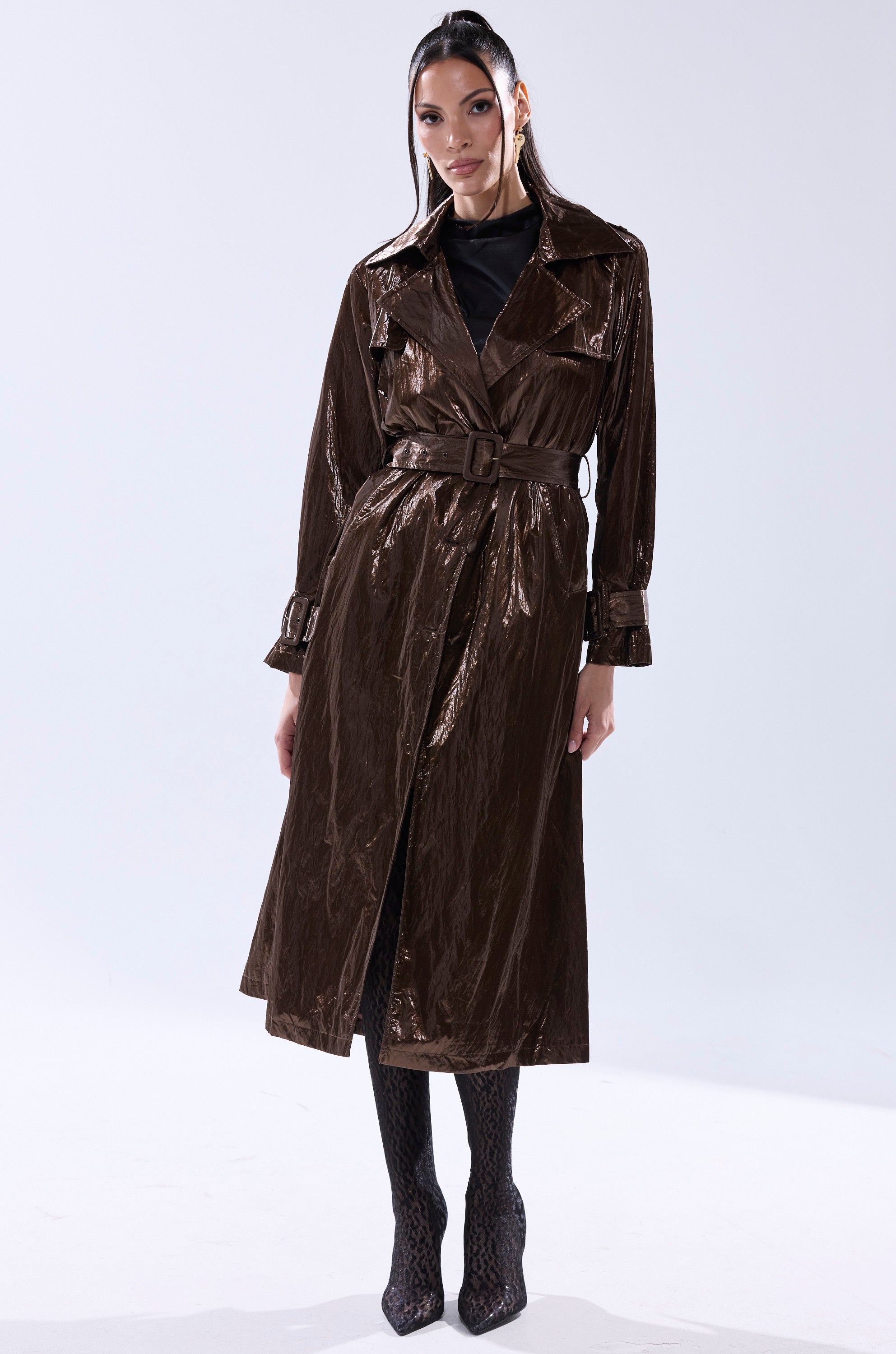 A woman stands facing forward wearing the WORK OF ART METALLIC TRENCH, a bronze longline coat with a belted waist, styled with a black top, patterned tights, and black heels against a plain light background. 7