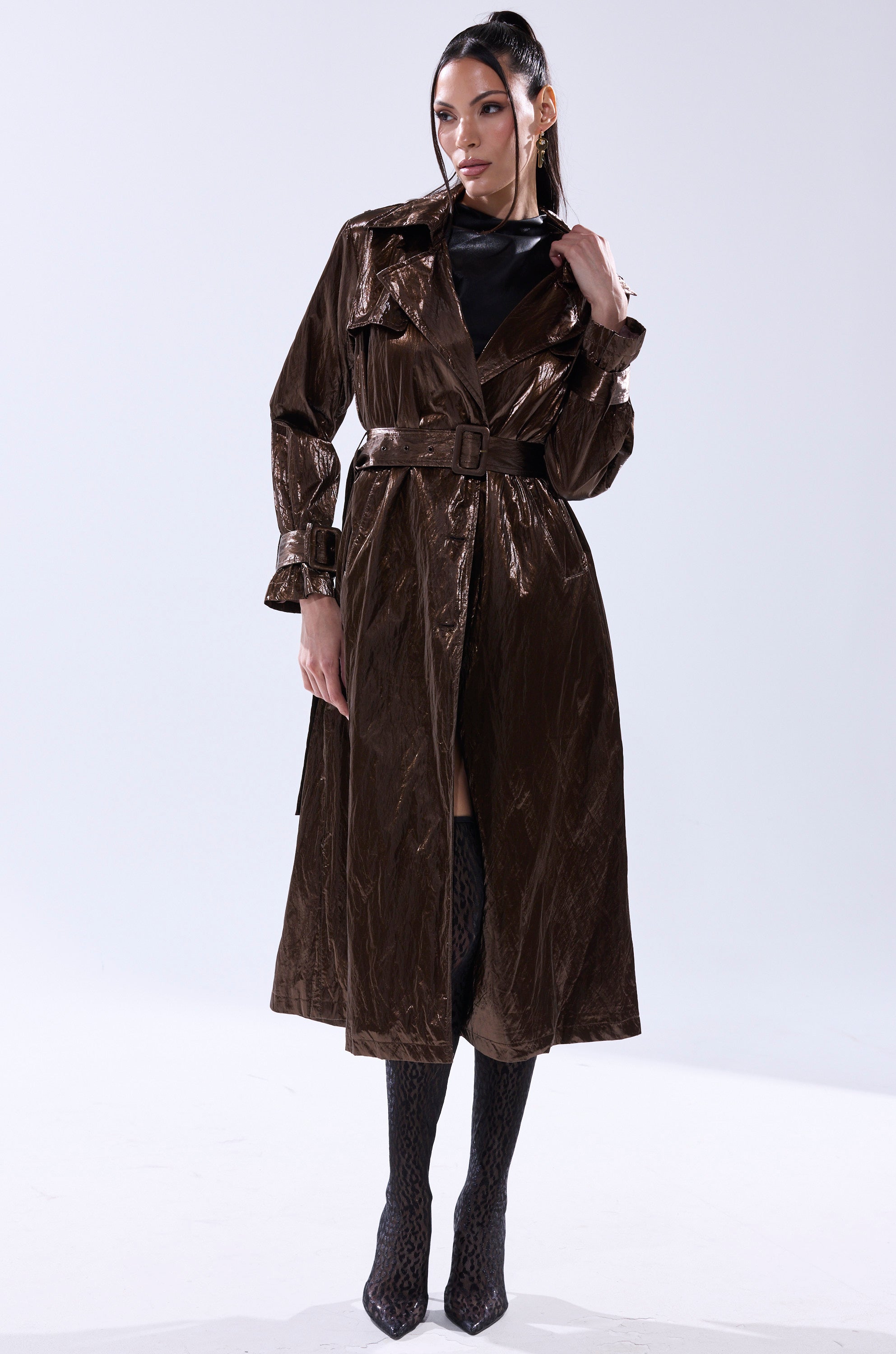A woman with long dark hair strikes a confident pose against a plain background, wearing the WORK OF ART METALLIC TRENCH in bronze over a black outfit, paired with knee-high patterned boots. 8