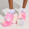 A person dons the GIRLY POP LACE RUFFLE SOCKS IN WHITE, paired with fluffy platform slippers showcasing a vibrant mix of pink, white, and light blue hues against a simple light backdrop—a perfect fusion of whimsy and style.