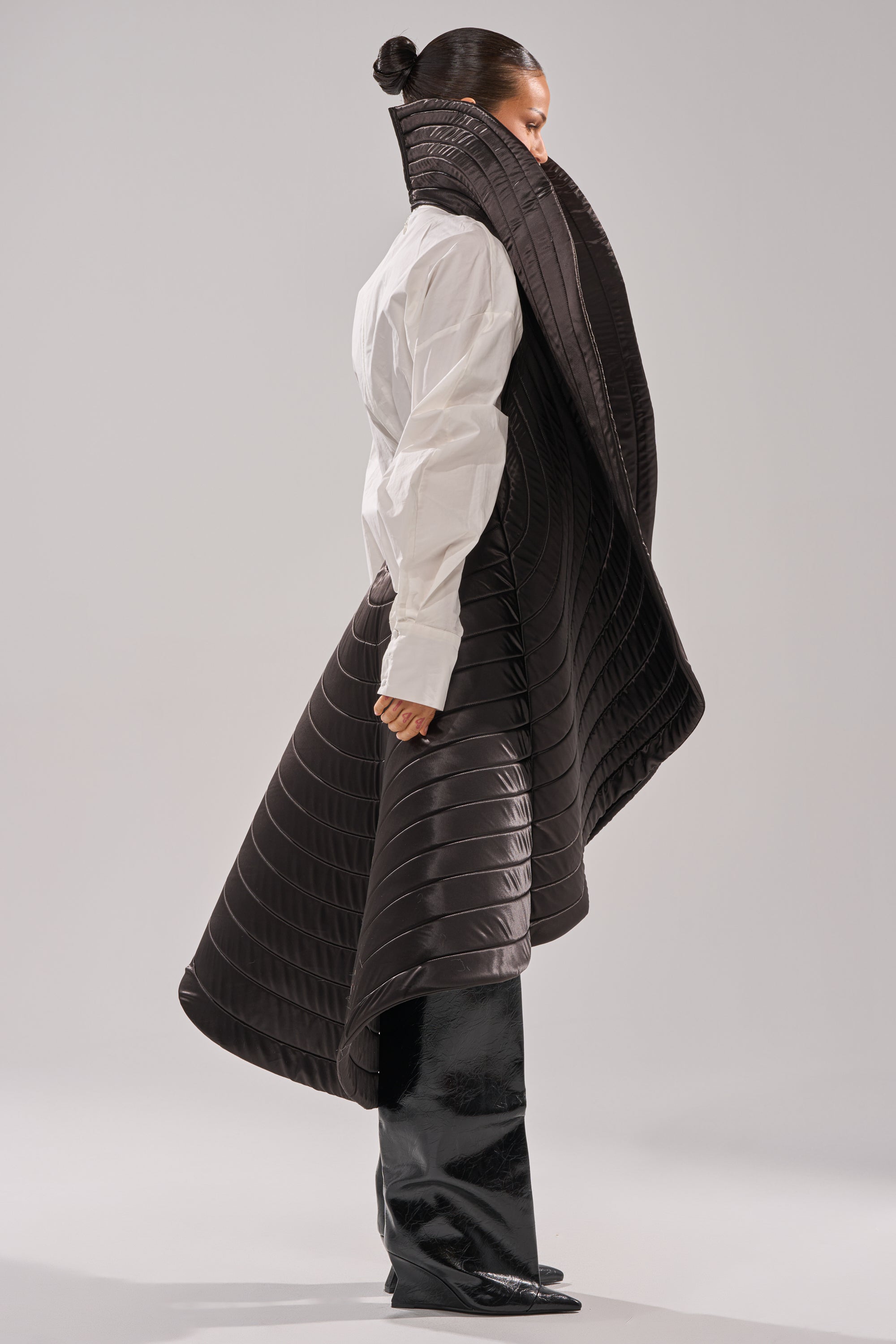 A person in profile wears the FOR THE DRAMA HIGH LOW VEST—an avant-garde, oversized quilted black vest with a tall collar—layered over a white shirt and shiny black boots, set against a plain background.