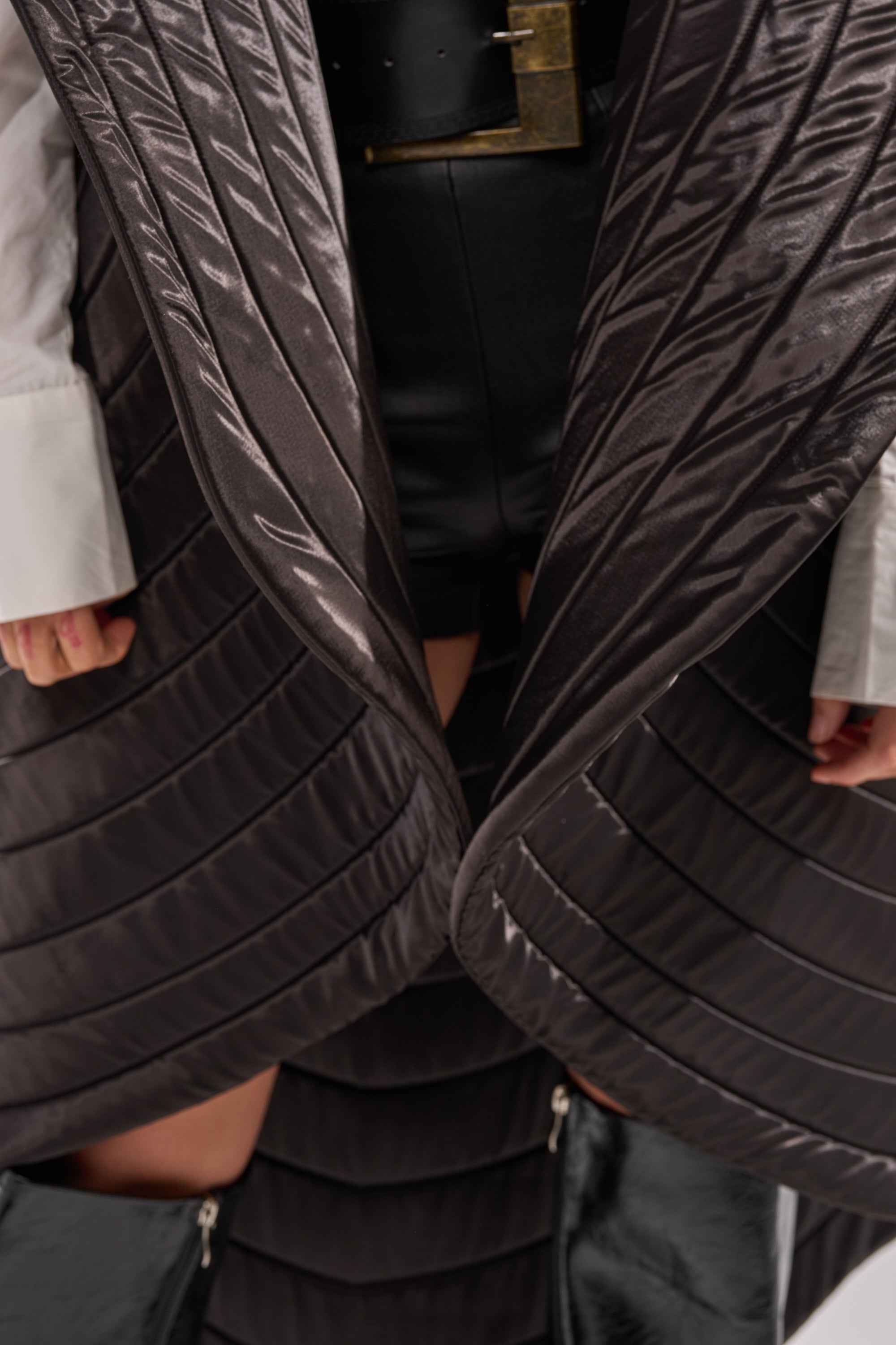 A person wears the shiny, quilted FOR THE DRAMA HIGH LOW VEST with a sculptural collar over a white shirt and black shorts, paired with black knee-high boots. Only the lower torso and upper legs are shown.