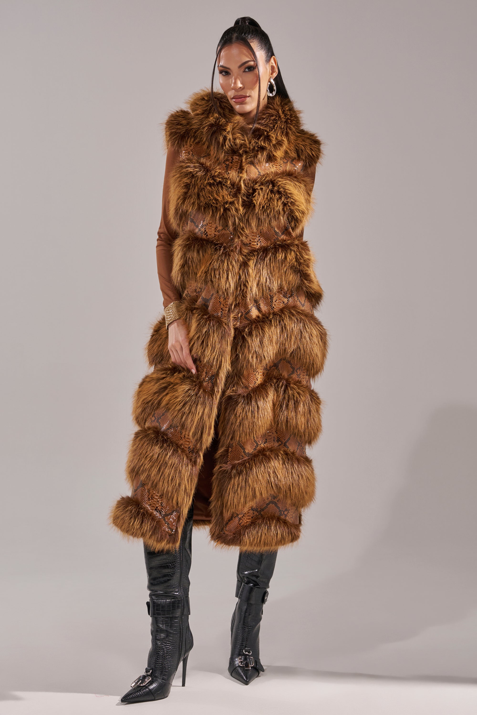 A woman with long straight hair and hoop earrings stands against a gray background, exuding chic winter style in the GOOD OMEN FUR TRIM VEST over a brown outfit and black buckle knee-high boots. 0