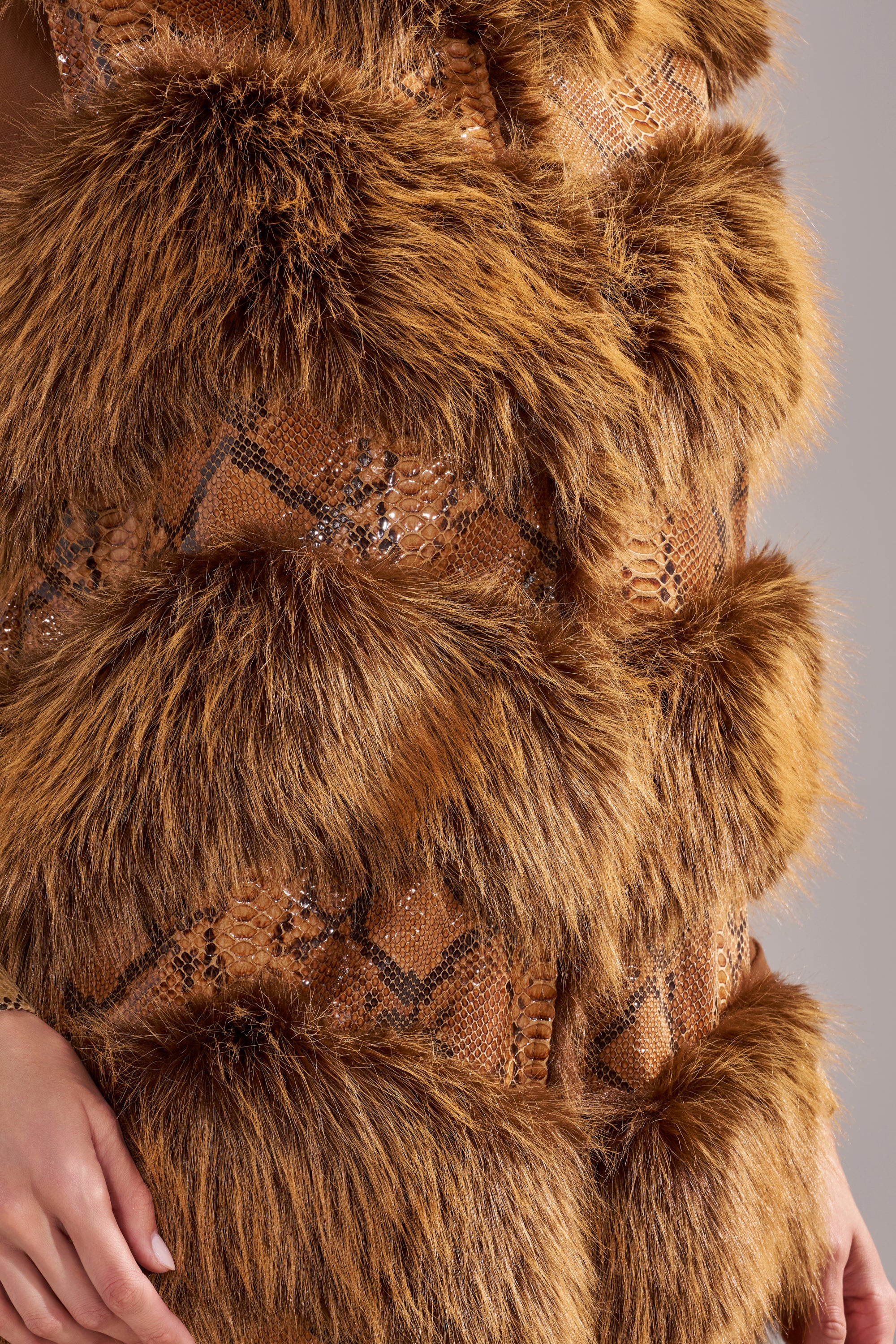 Close-up of the GOOD OMEN FUR TRIM VEST, showcasing alternating bands of brown faux fur and textured snake-print fabric; a hand is partially visible resting at the bottom edge on the left. 11