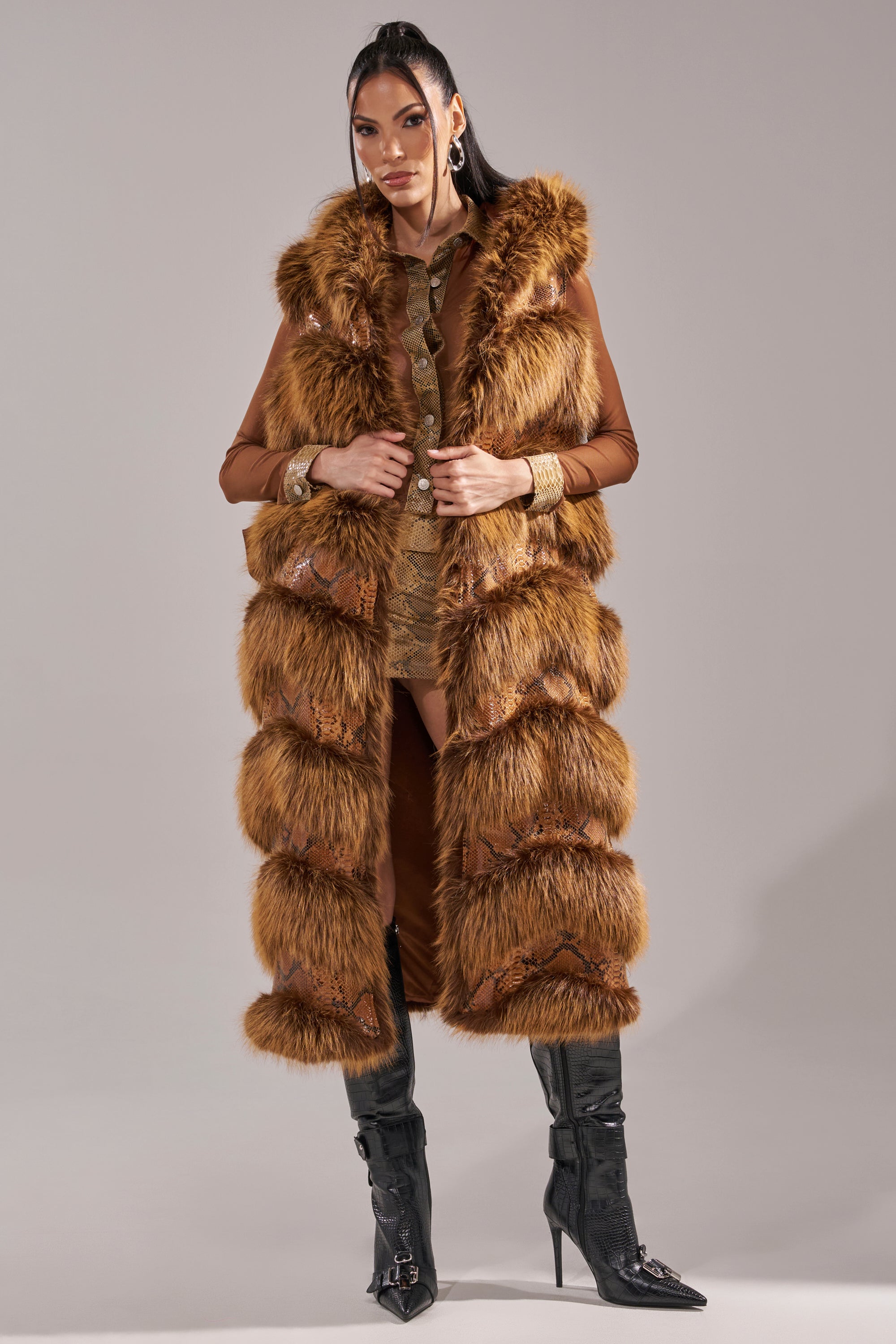 A woman stands confidently in the GOOD OMEN FUR TRIM VEST over a brown outfit with black knee-high boots. She has long dark hair, bold earrings, and bracelets, posing against a plain light gray background. 3