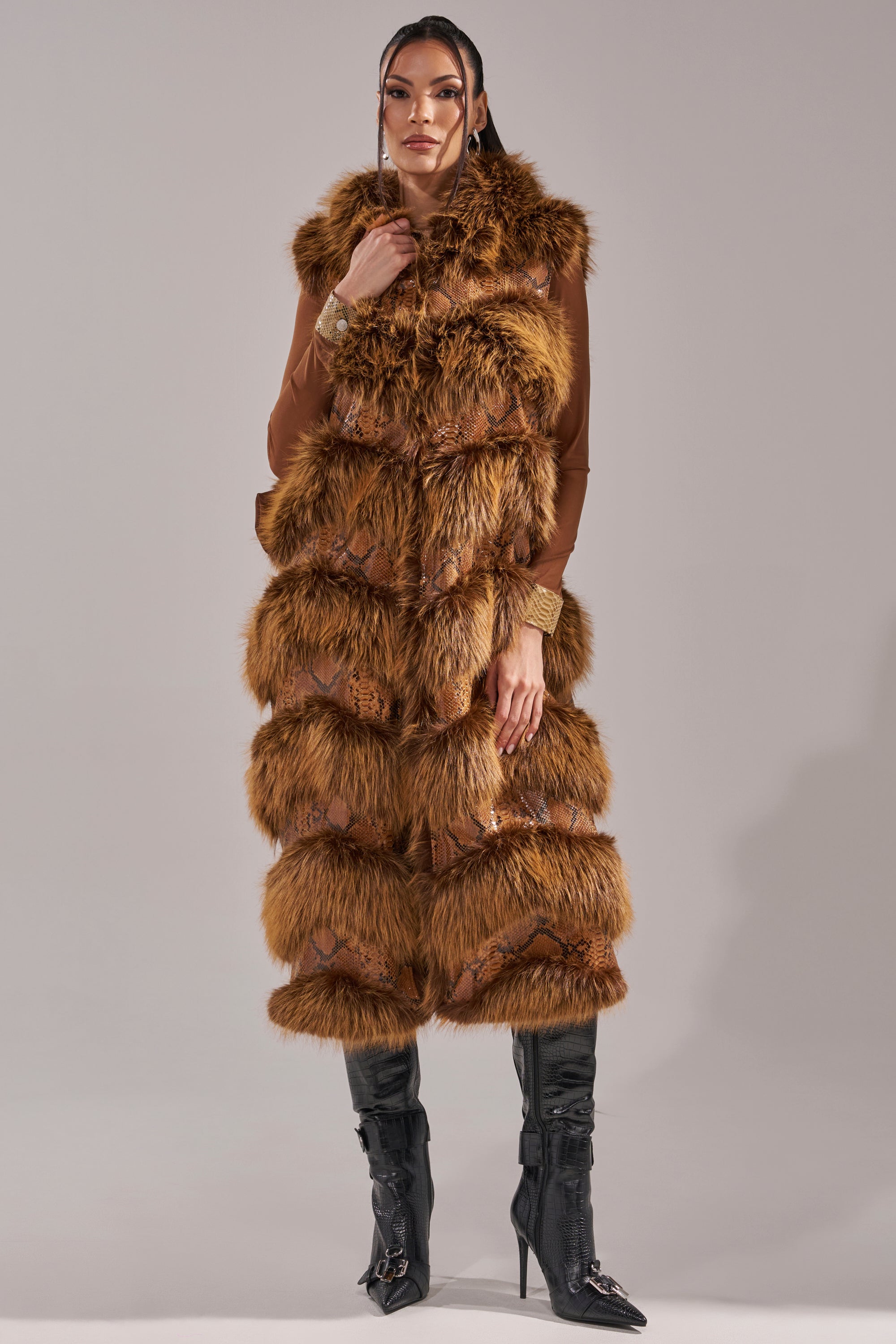 A woman poses against a gray backdrop wearing the GOOD OMEN FUR TRIM VEST over a fitted brown top and black knee-high boots, her hair neatly styled and one hand touching her collar, showcasing subtle snakeskin print accents. 6