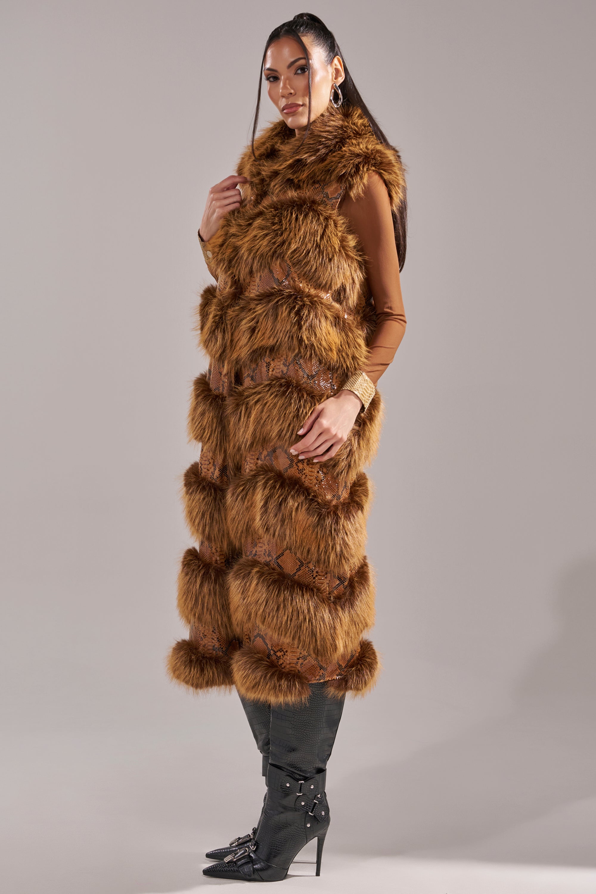 A woman models the GOOD OMEN FUR TRIM VEST, a long brown faux fur piece layered over a fitted top and styled with black knee-high boots with decorative accents. She poses sideways, exuding confidence against a plain backdrop. 7