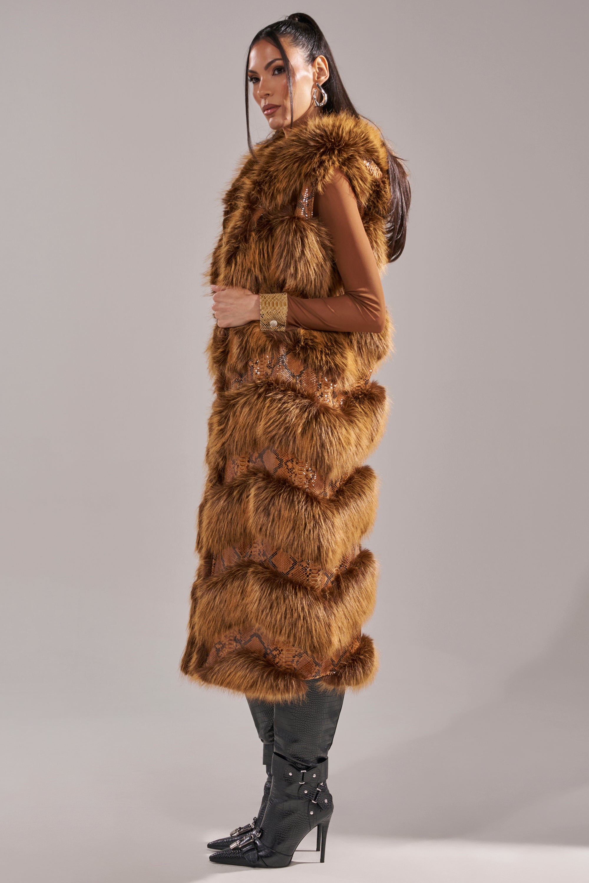 A woman in profile wears the GOOD OMEN FUR TRIM VEST, a long brown faux fur vest, over a brown top with black knee-high boots and gold hoop earrings, set against a plain gray background. 8