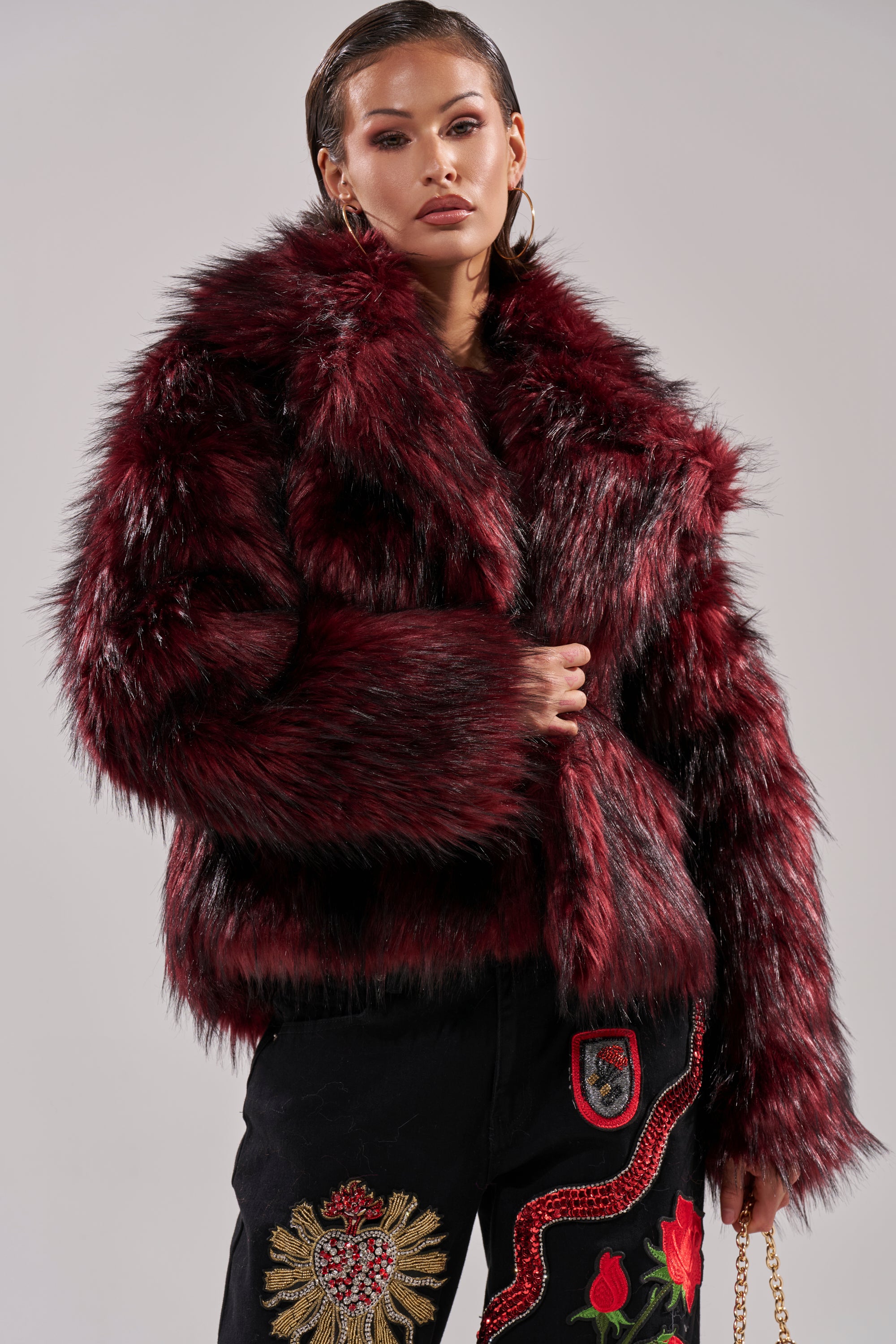 A woman models the MICHIGAN AVE FAUX FUR red jacket and black pants with gold crest and red rose embroidery, standing confidently against a light gray background.