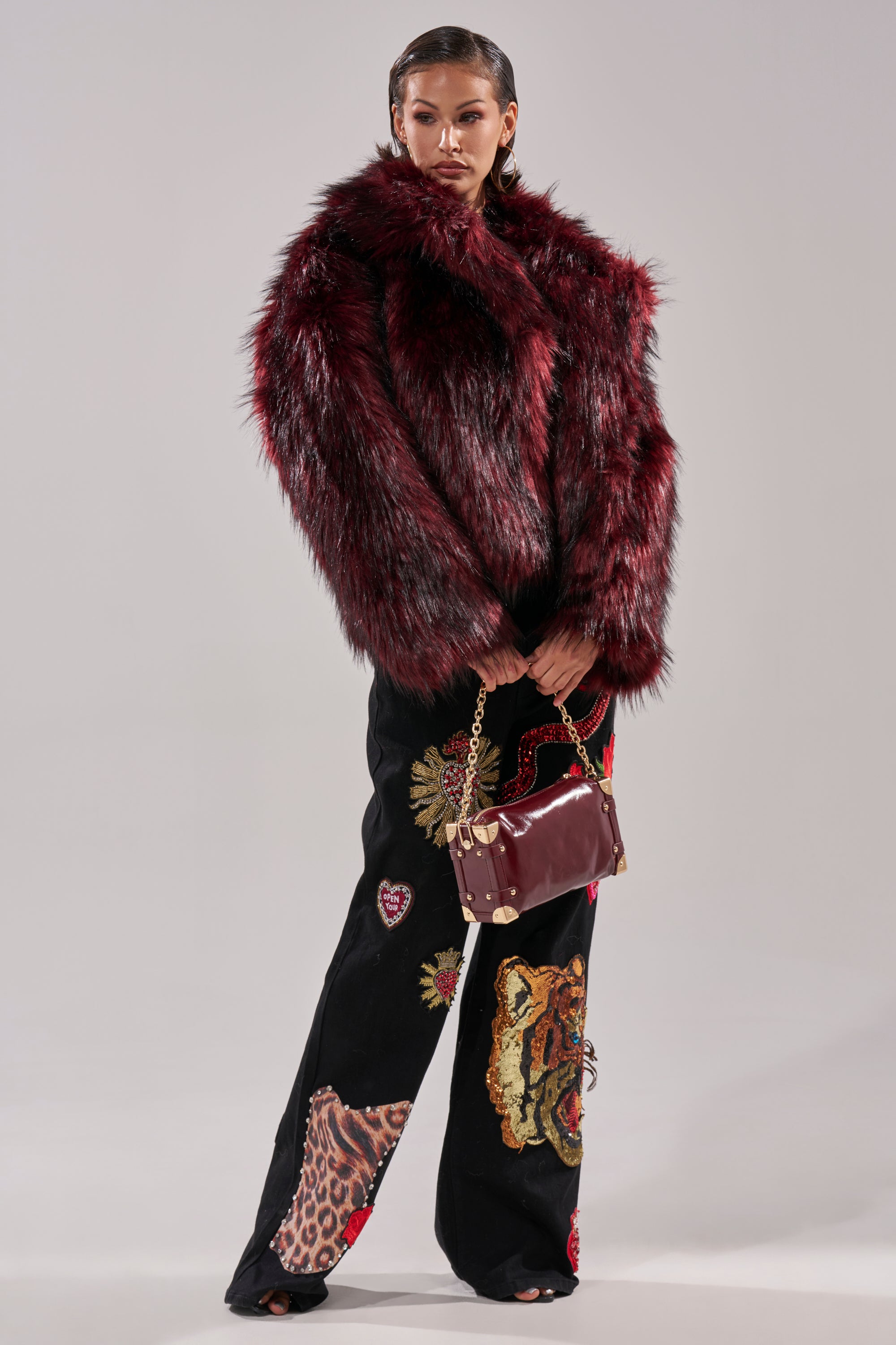 A person stands confidently in the luxurious MICHIGAN AVE FAUX FUR jacket, paired with black pants featuring embroidered animal designs, and holds a red handbag with a gold chain strap. The bold red faux fur stands out against the light background.