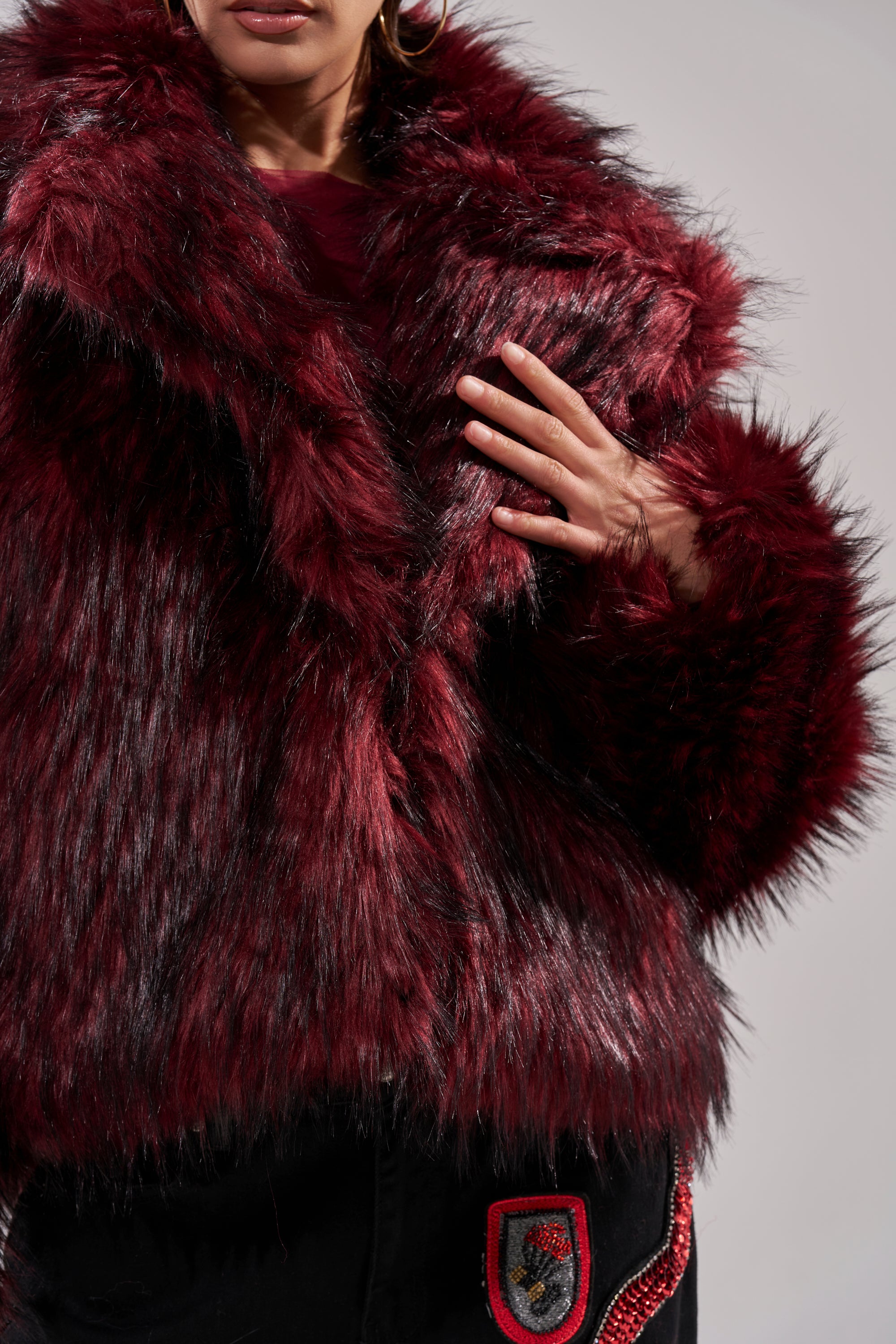 Clad in the luxurious MICHIGAN AVE FAUX FUR jacket, a person displays only their lower face and hand against a plain, light background. The jacket features dark red faux fur with a thick, fluffy texture.