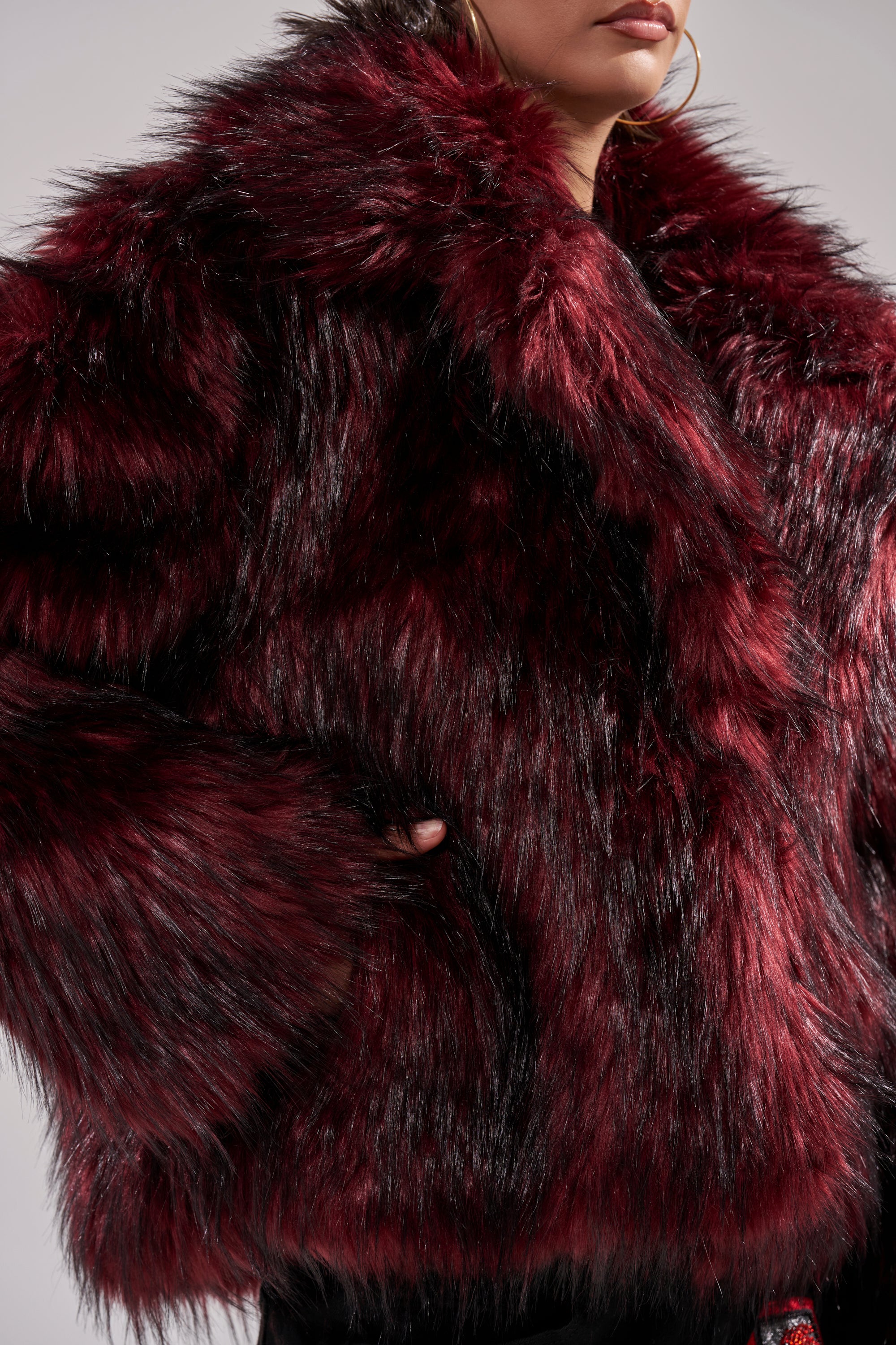 A person is shown from shoulders to hips wearing the MICHIGAN AVE FAUX FUR, a luxurious deep burgundy red jacket with black accents, plush thick texture, and their hand resting in one of the winter jacket’s pockets.