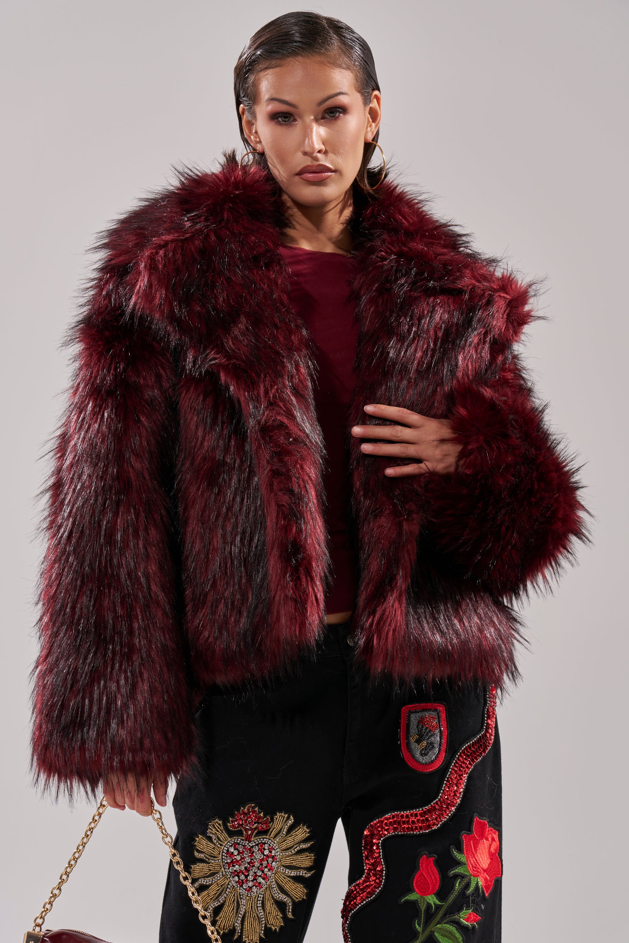 A woman embraces winter chic in the MICHIGAN AVE FAUX FUR, a deep red jacket over a matching top, paired with black pants with vibrant embroidery. She carries a gold chain purse and wears sleek, wet-look hair.