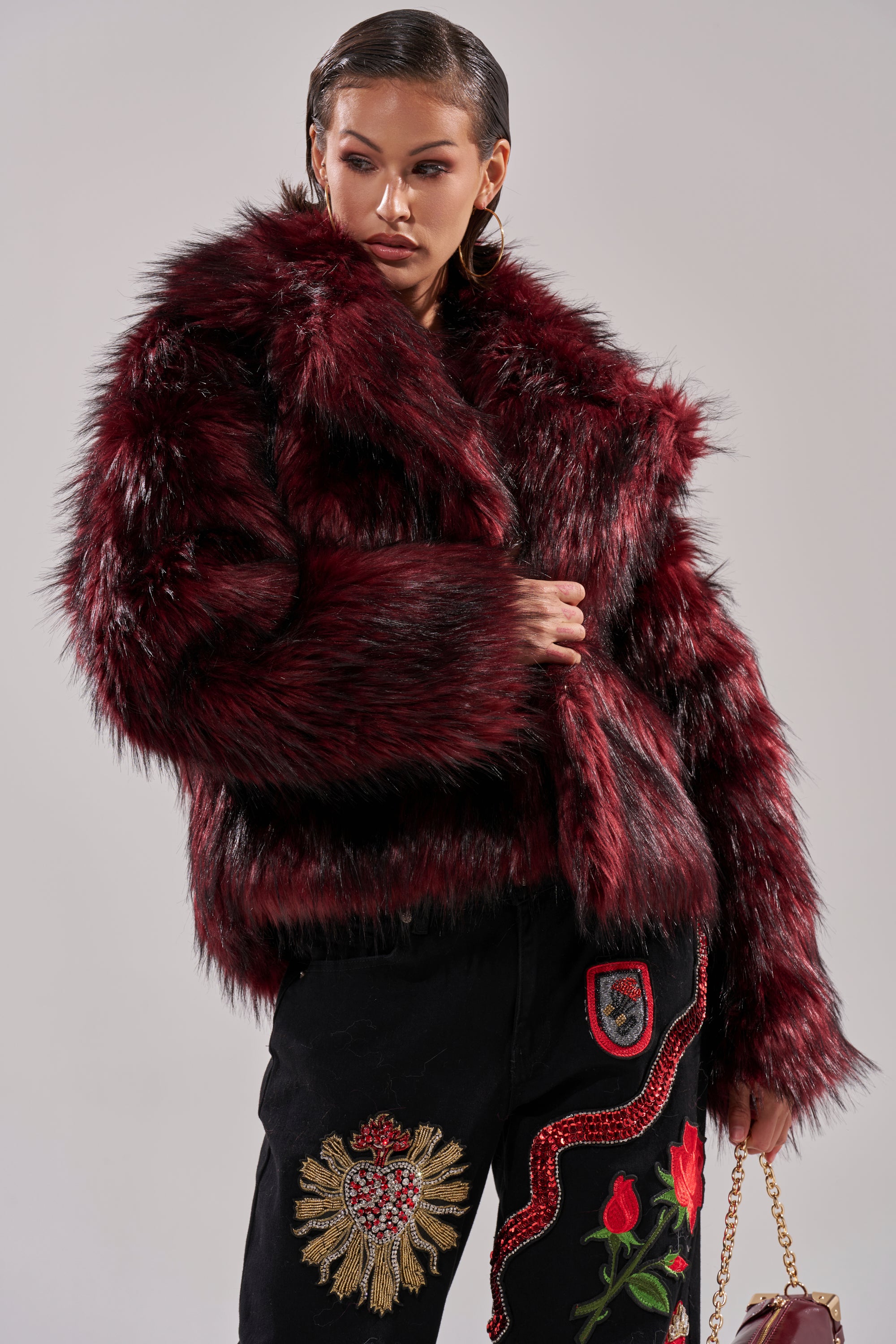 A woman in the MICHIGAN AVE FAUX FUR jacket and black embroidered pants holds a small handbag. With slicked-back hair and gold hoop earrings, she makes a bold winter-chic statement.