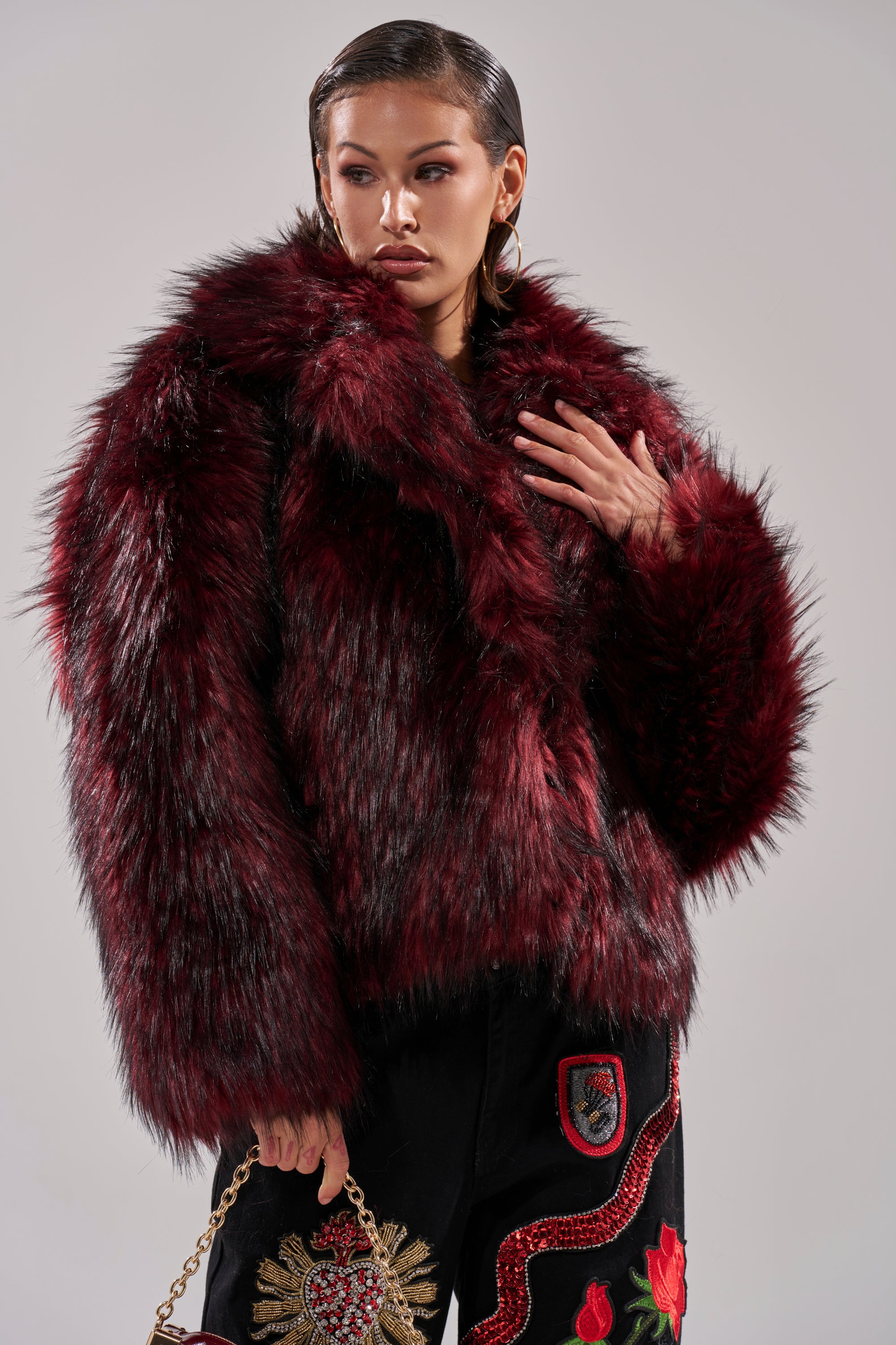 A woman wears the winter-chic MICHIGAN AVE FAUX FUR coat in luxe red over black embroidered pants, holding a gold chain handbag against a plain light background.