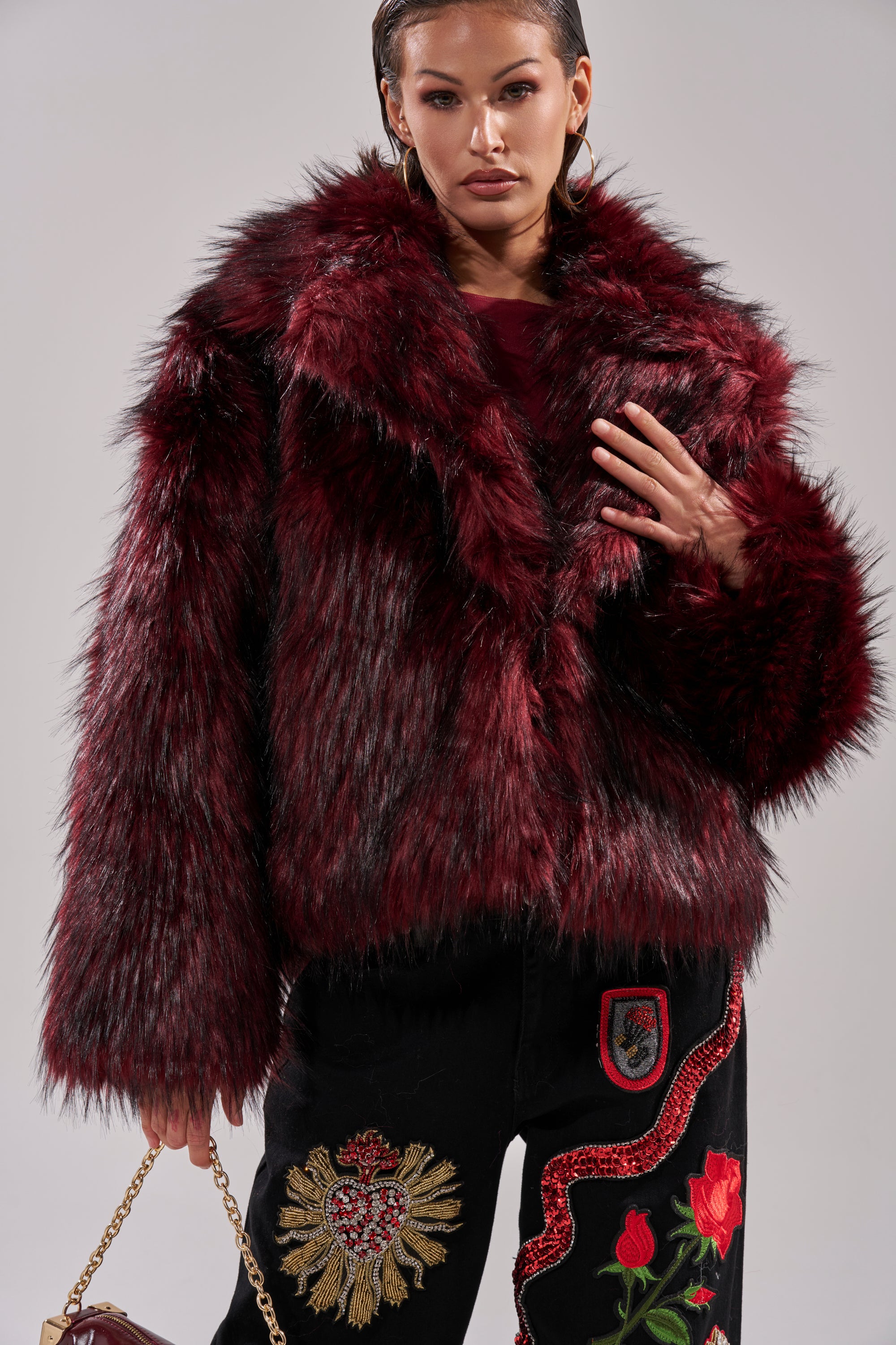 A woman poses against a plain background wearing the MICHIGAN AVE FAUX FUR jacket in red and black pants with gold and red embroidery, holding a small handbag.