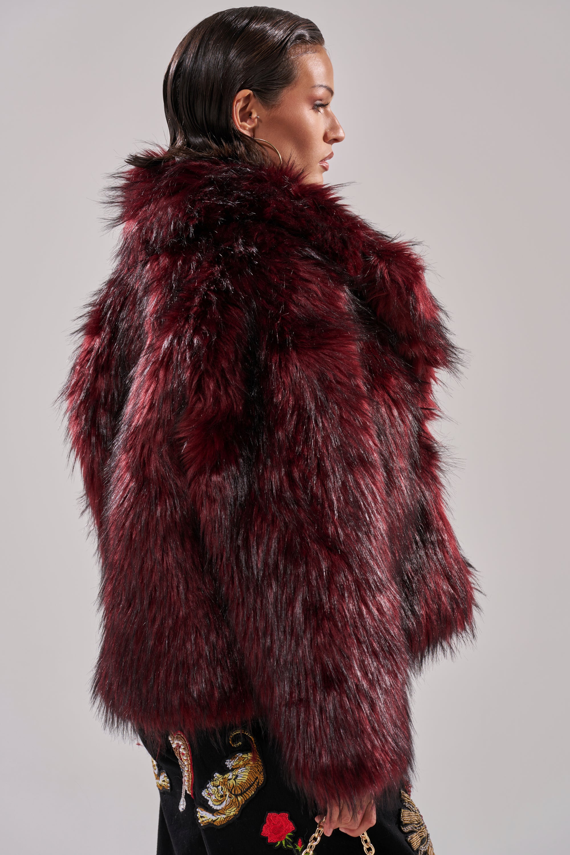 Wearing the MICHIGAN AVE FAUX FUR in deep burgundy, a person with slicked-back dark hair pairs this winter-chic jacket with black embroidered pants, standing in profile against a plain background.