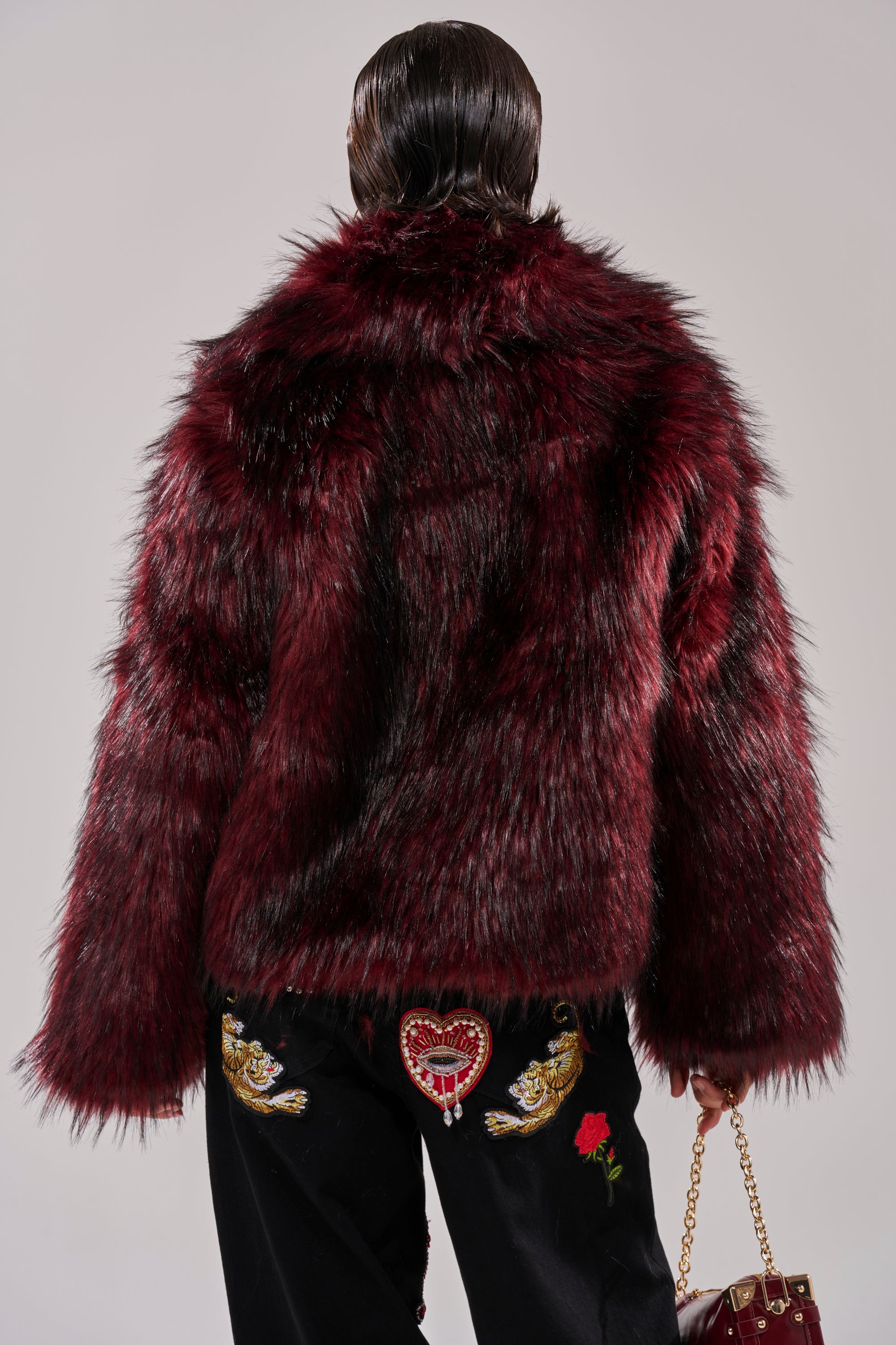 A person with slicked-back hair is seen from behind in the winter-chic MICHIGAN AVE FAUX FUR red coat and black pants with embroidered designs, including a red heart and gold accents, holding a matching red bag.