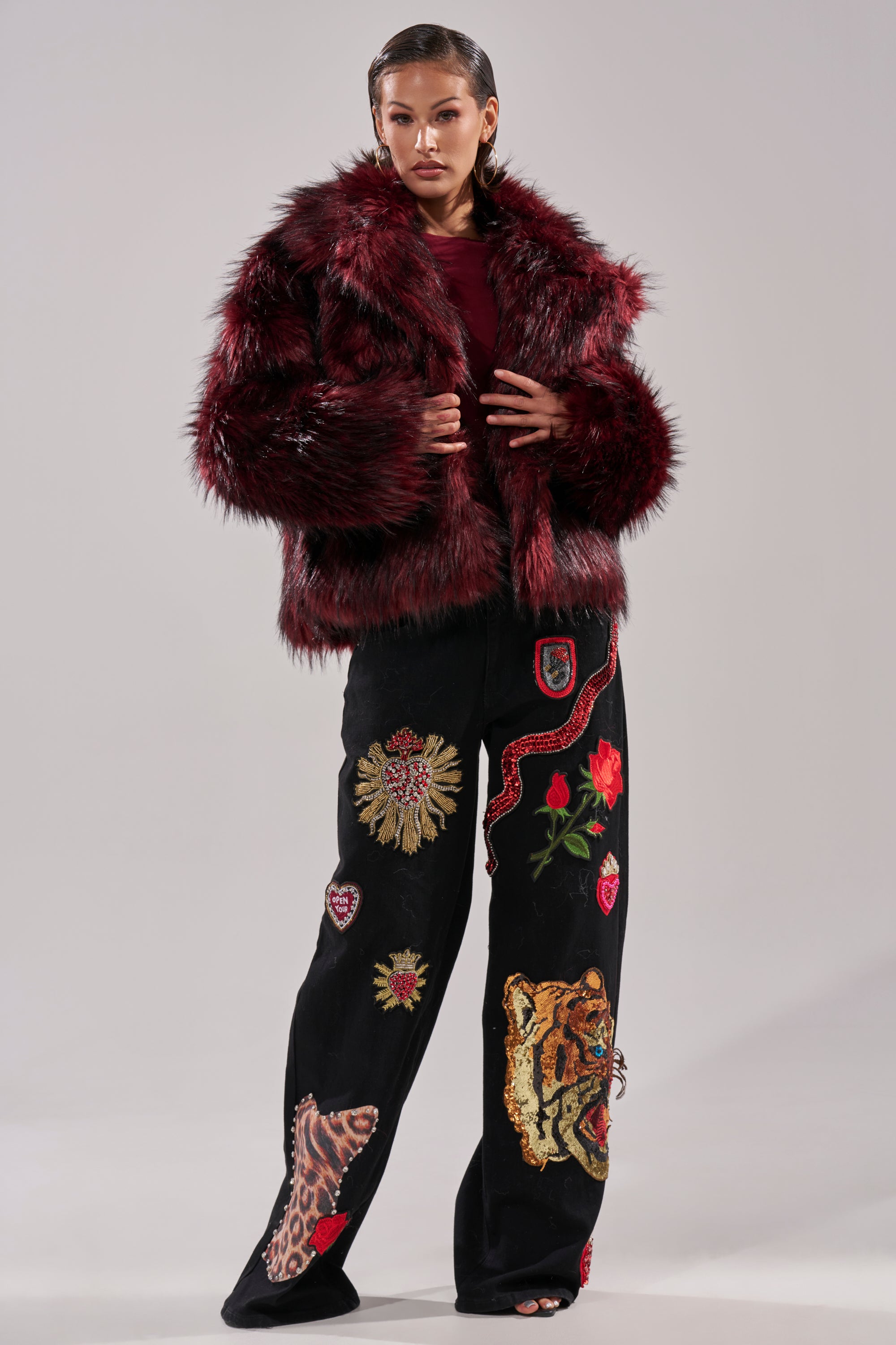 A person stands confidently in the MICHIGAN AVE FAUX FUR deep red jacket and black wide-leg pants with tiger and rose embroidery, set against a simple light background.