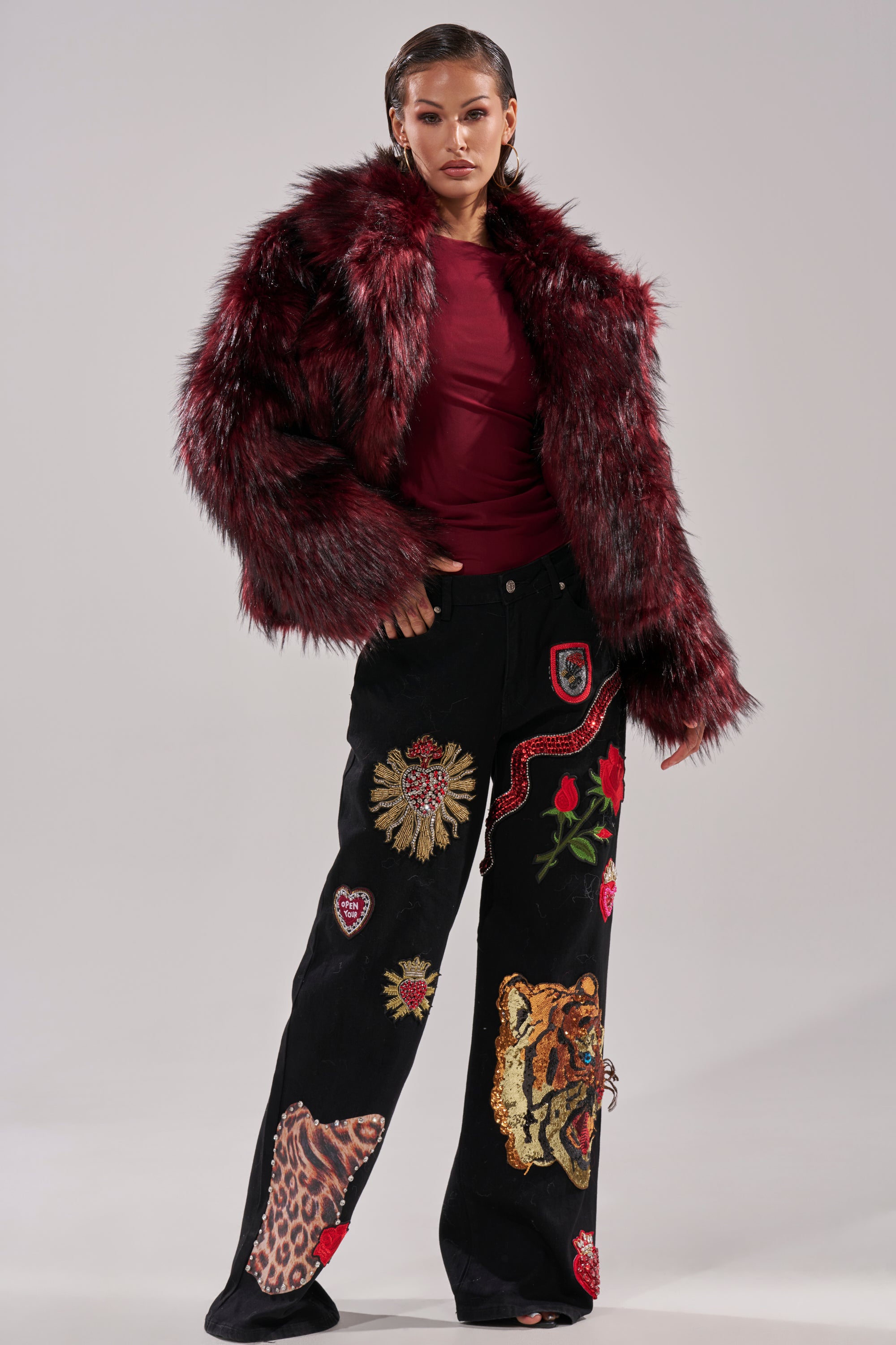 Woman models the MICHIGAN AVE FAUX FUR jacket in red, layered over a dark red top and black pants with colorful animal and floral patches, set against a plain light background.