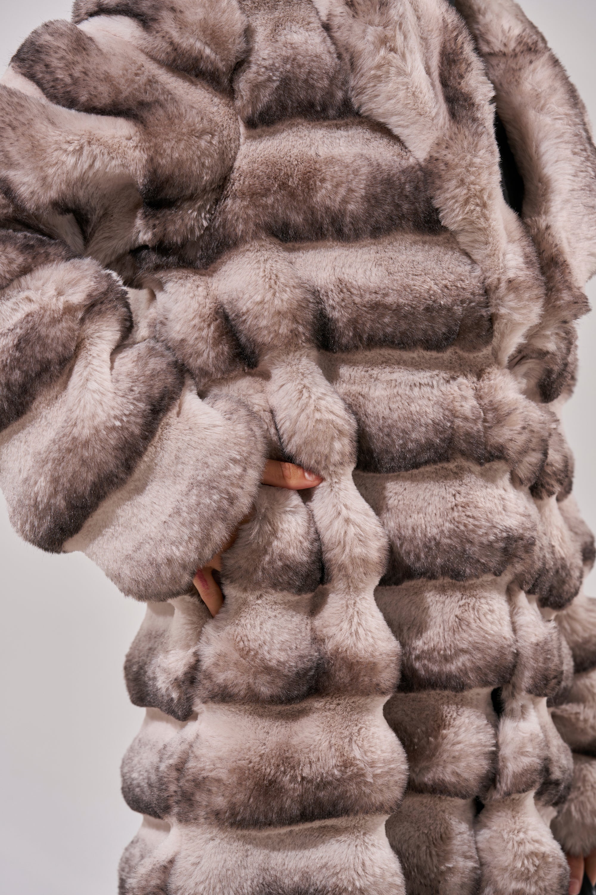 A person wearing the FAIRBANKS MAXI FAUX FUR, a thick, plush striped longline coat in beige and brown shades, with one hand tucked into the side pocket.