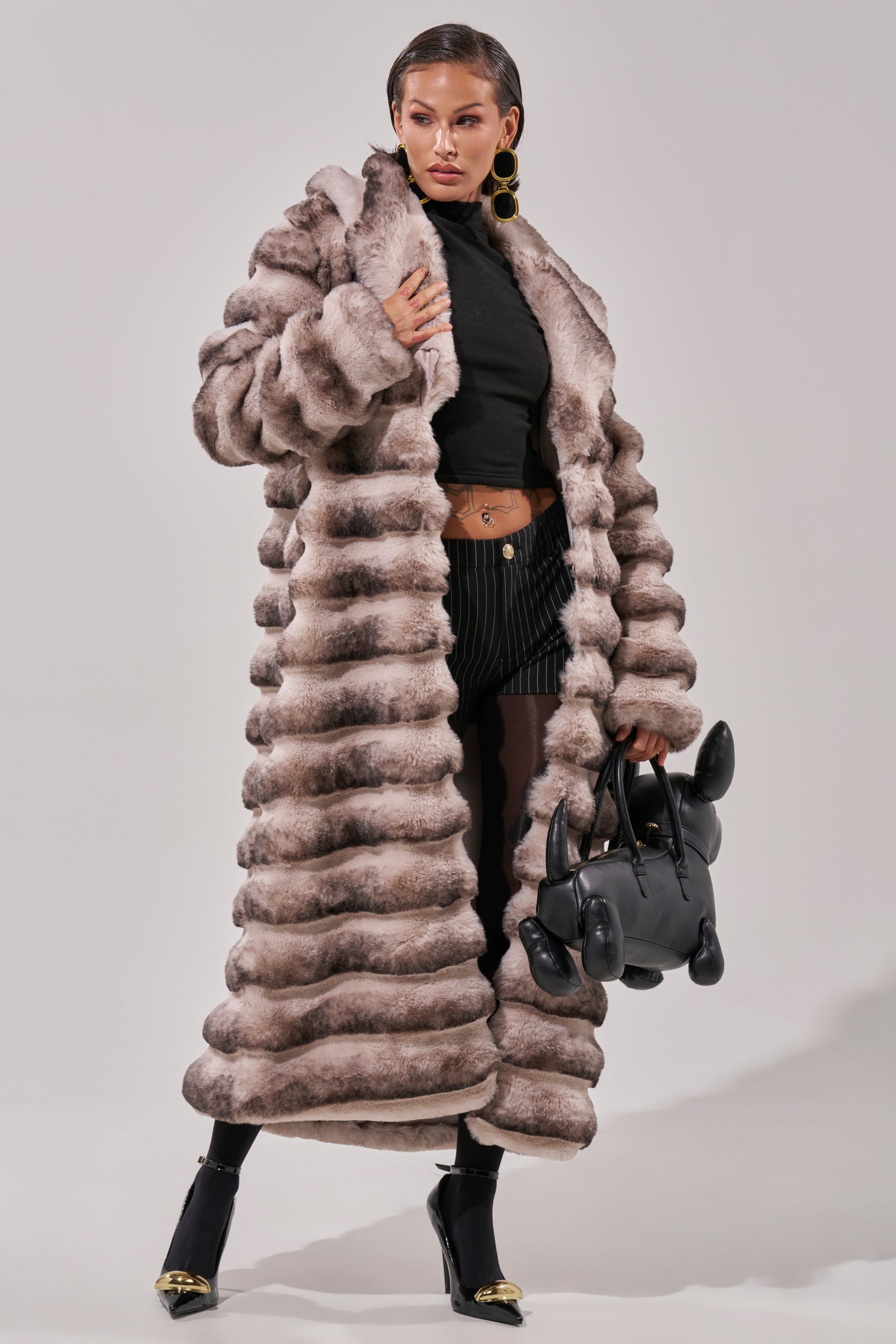 A woman in the FAIRBANKS MAXI FAUX FUR coat, black turtleneck crop top, pinstriped shorts, black tights, and gold-accented black heels holds a whimsical balloon dog-shaped black handbag.