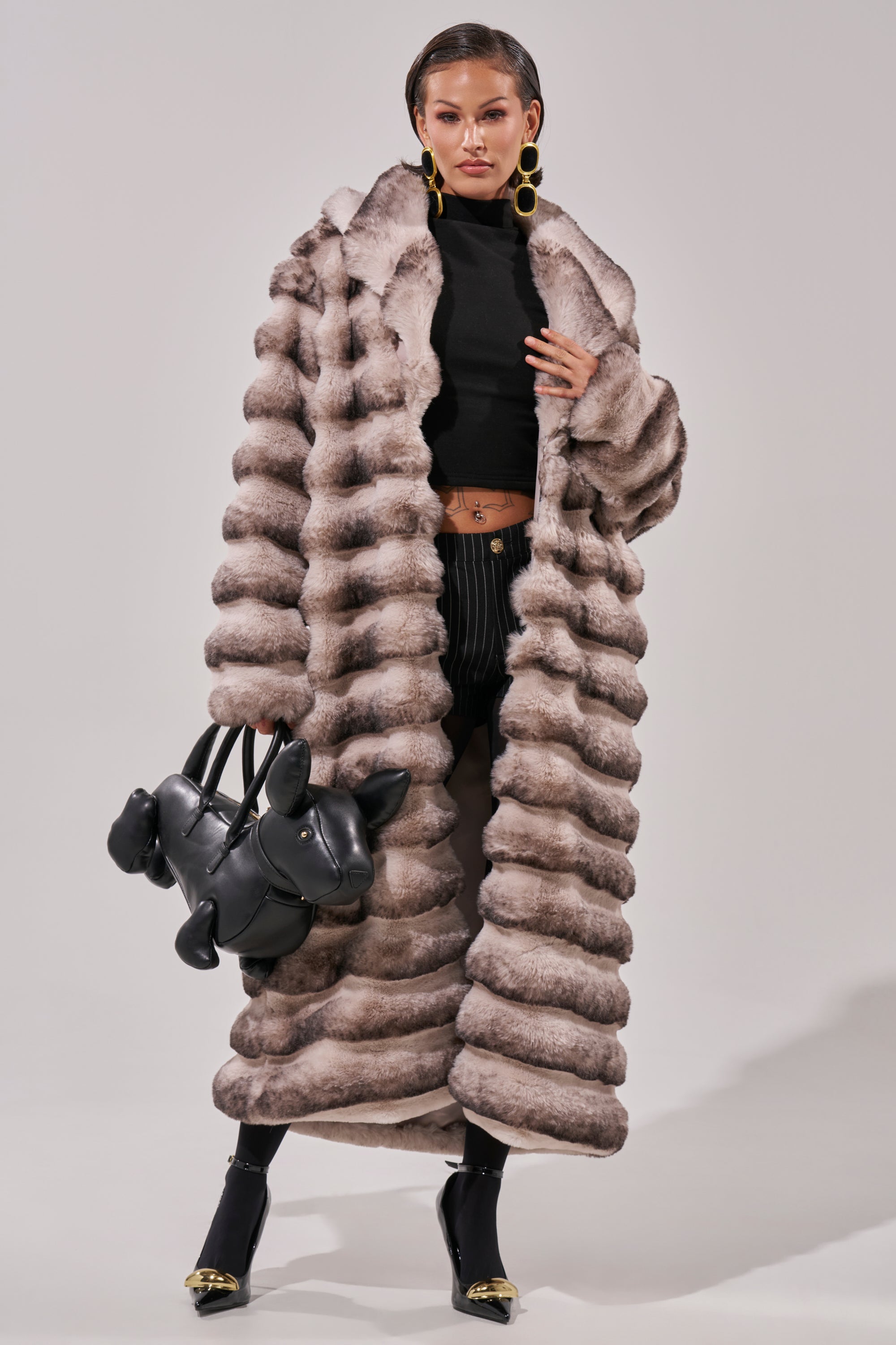 A woman poses in the FAIRBANKS MAXI FAUX FUR coat with a black turtleneck crop top, striped pants, heeled boots, a black balloon dog handbag, and large round gold statement earrings.