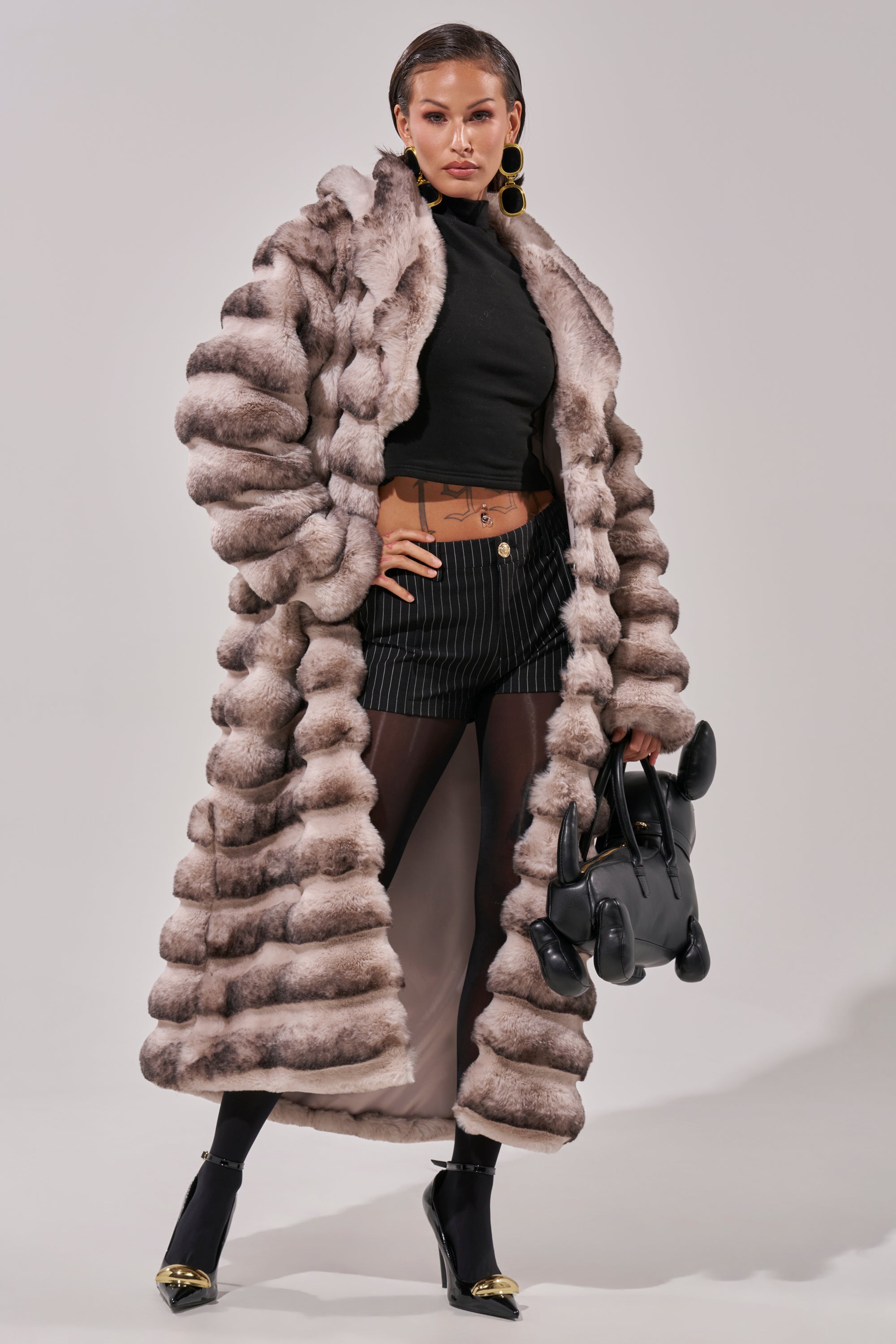 Wearing the FAIRBANKS MAXI FAUX FUR jacket, a woman in a black turtleneck, pinstripe shorts, sheer tights, and gold-accented heels holds a black dog-shaped handbag and poses confidently against a plain background.