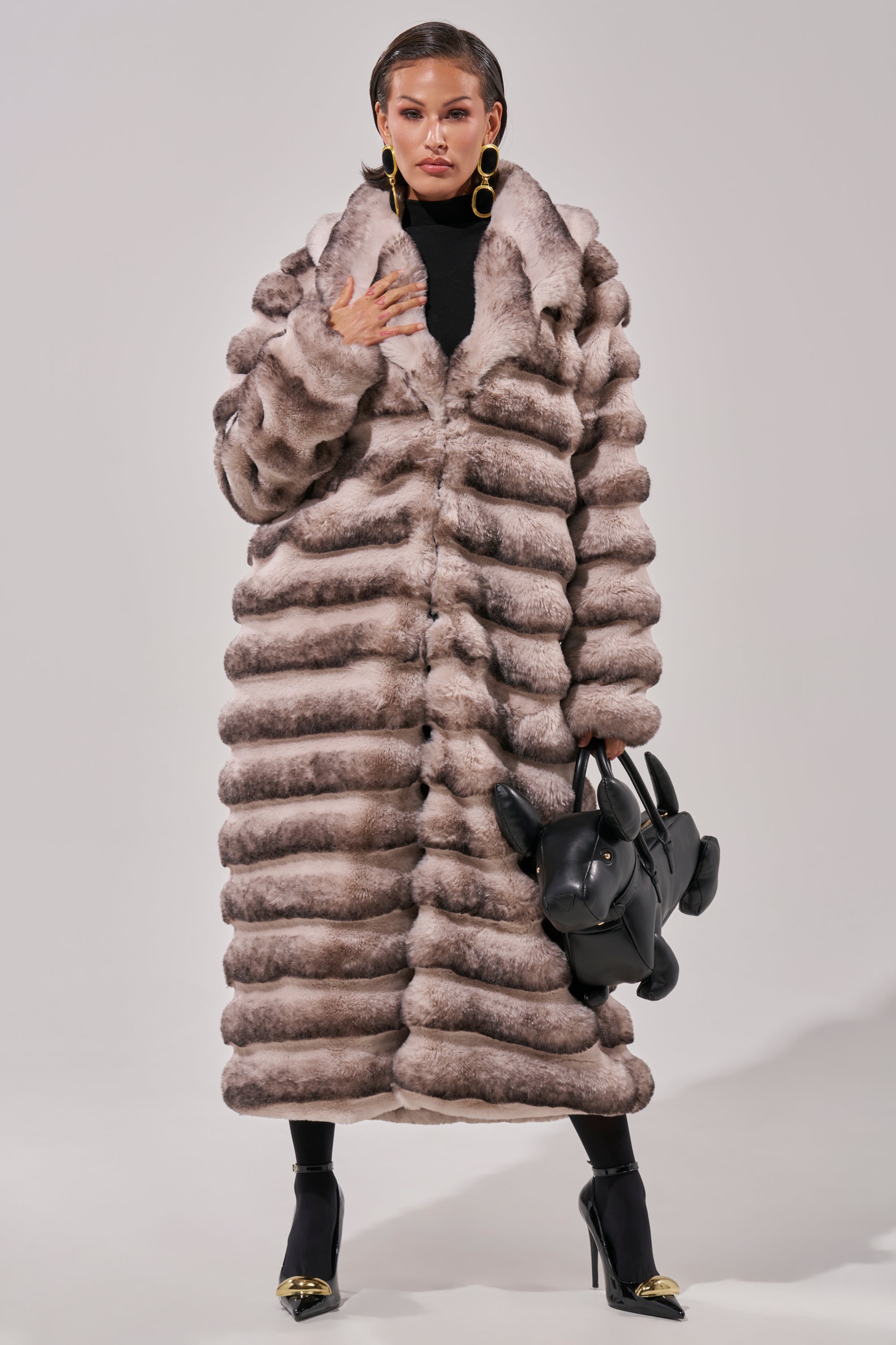 Wearing the FAIRBANKS MAXI FAUX FUR coat over a black turtleneck and tights with gold-tipped heels, she carries a playful dog-shaped handbag and completes her look with bold hoop earrings.