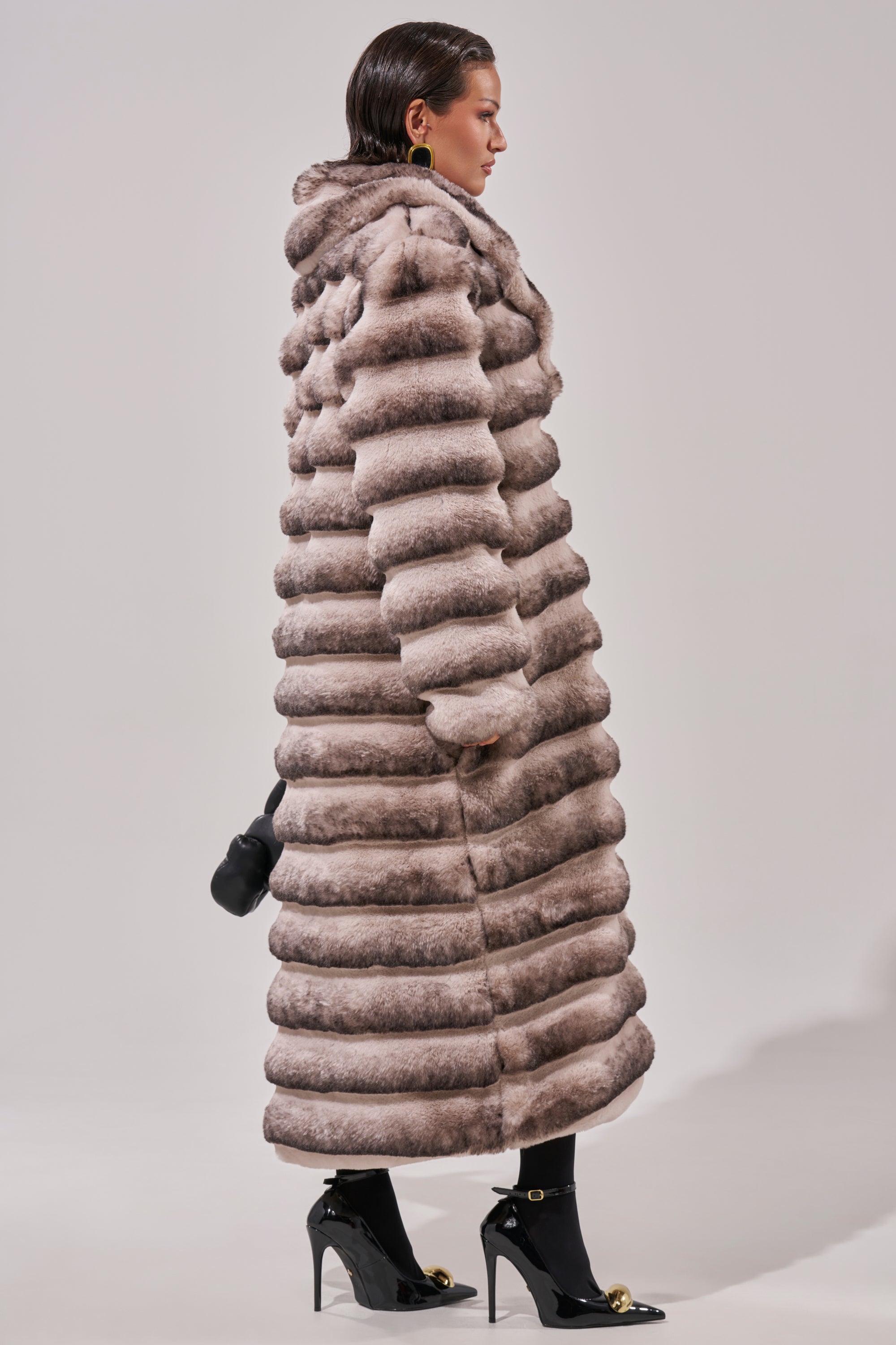 Wearing the FAIRBANKS MAXI FAUX FUR coat with a hood, black tights, and black heels with gold accents, she carries a small black clutch. Her slicked-back hair completes the polished look.