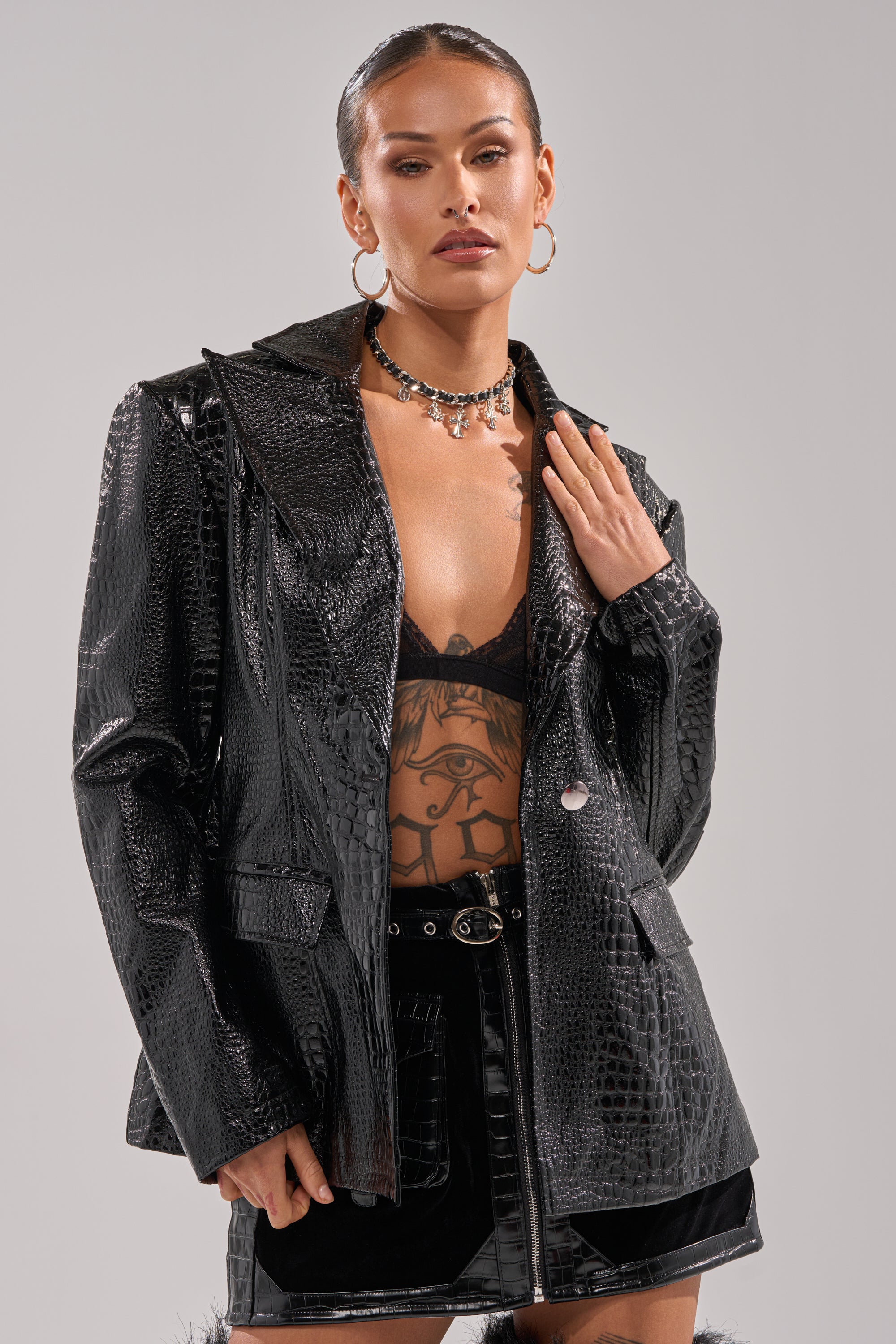 A woman with slicked-back hair wears the FOR THE DRAMA CROC BLAZER over a black bralette, revealing a chest tattoo. She pairs it with hoop earrings, a choker necklace, and a black mini skirt for a bold look.